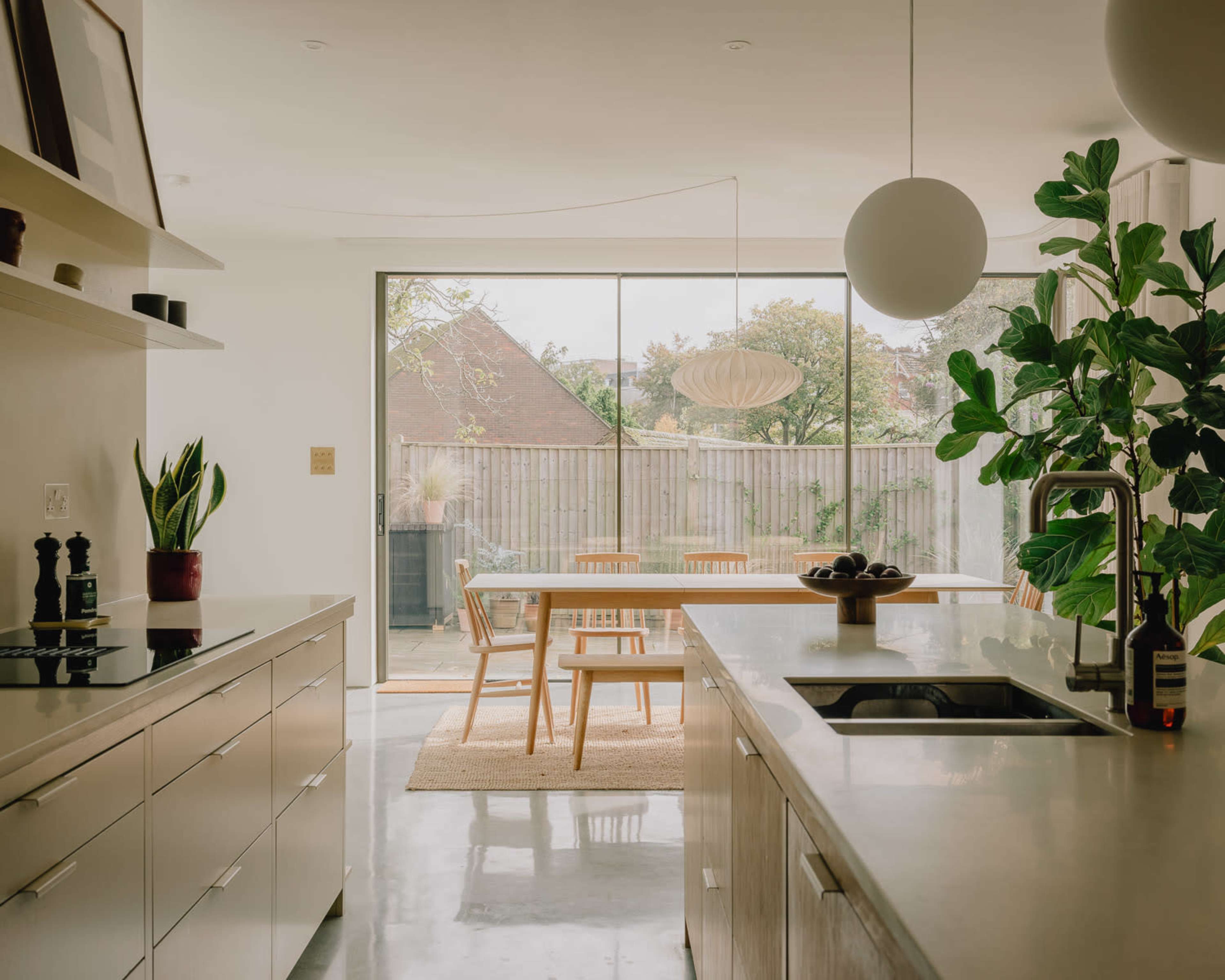 A modern kitchen features a long countertop, sleek cabinetry, and an open view of a dining area with wooden chairs and potted plants through large sliding glass doors.
