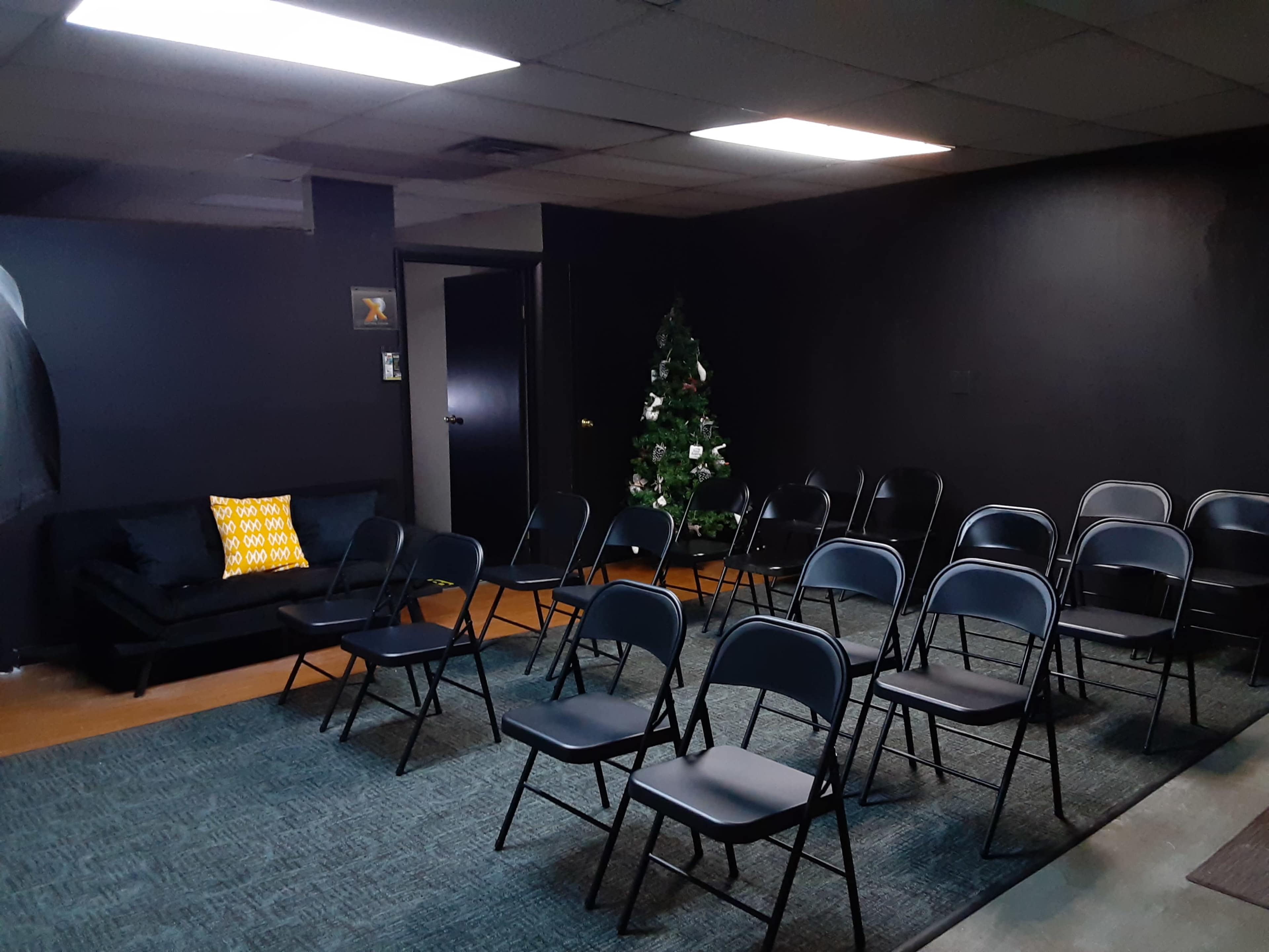 A sparsely furnished room with black walls, a few rows of folding chairs, and a decorated Christmas tree in the corner.