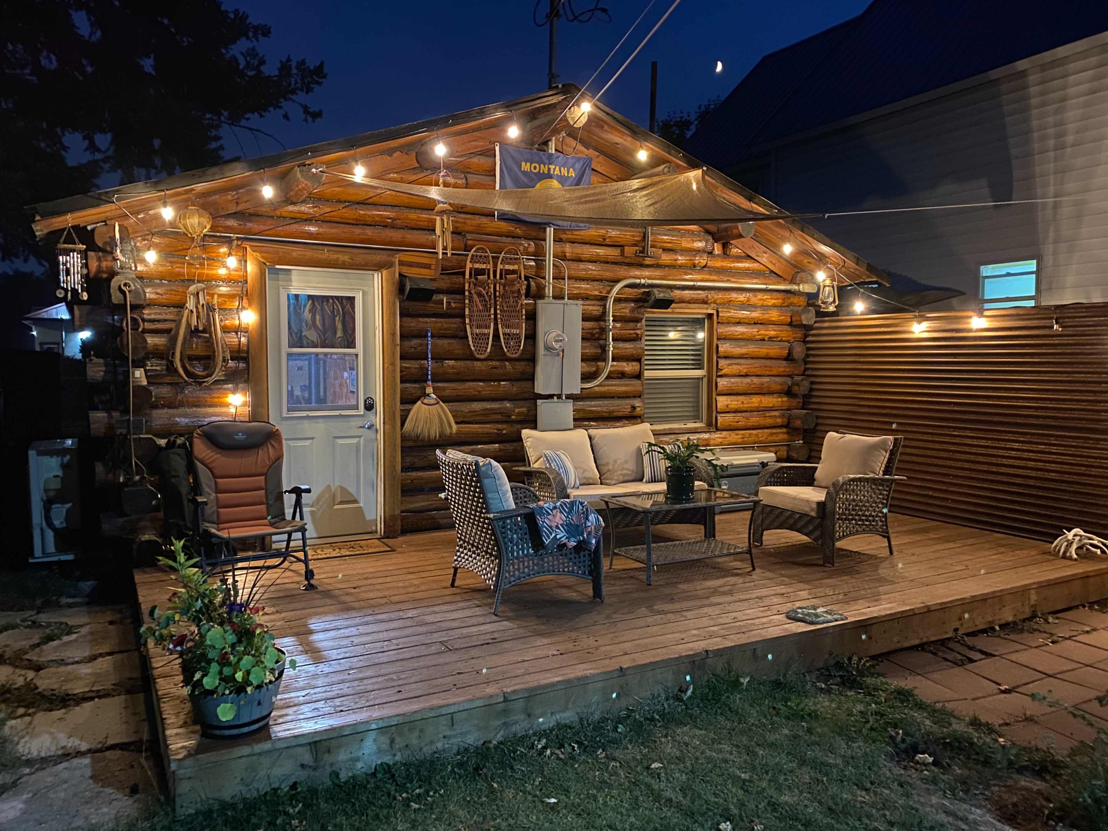 A log cabin with a wooden deck is illuminated by string lights, featuring outdoor furniture and potted plants.
