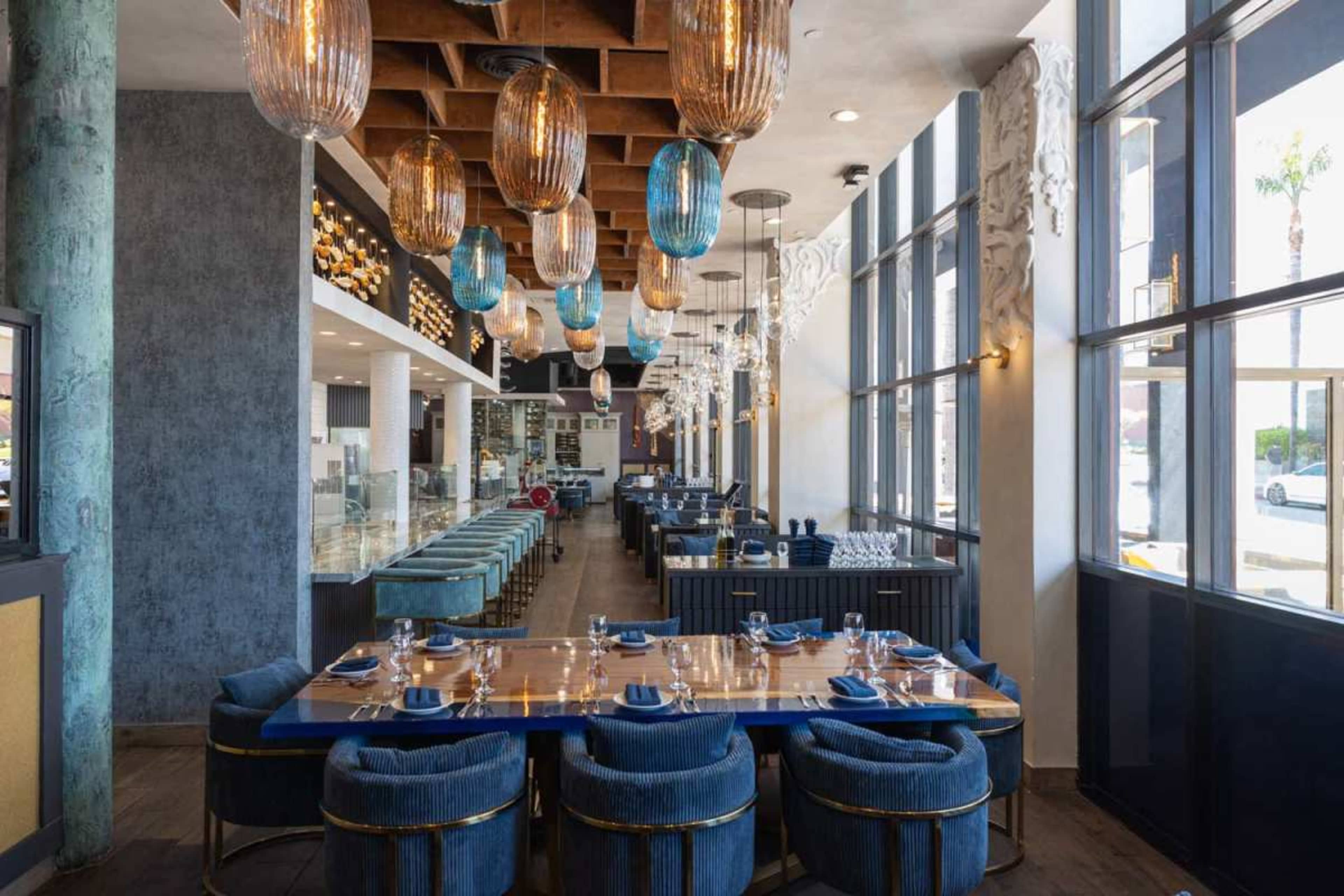 The image shows a modern restaurant interior with blue and gold accents, featuring a long wooden dining table surrounded by upholstered chairs and large windows allowing natural light to enter.
