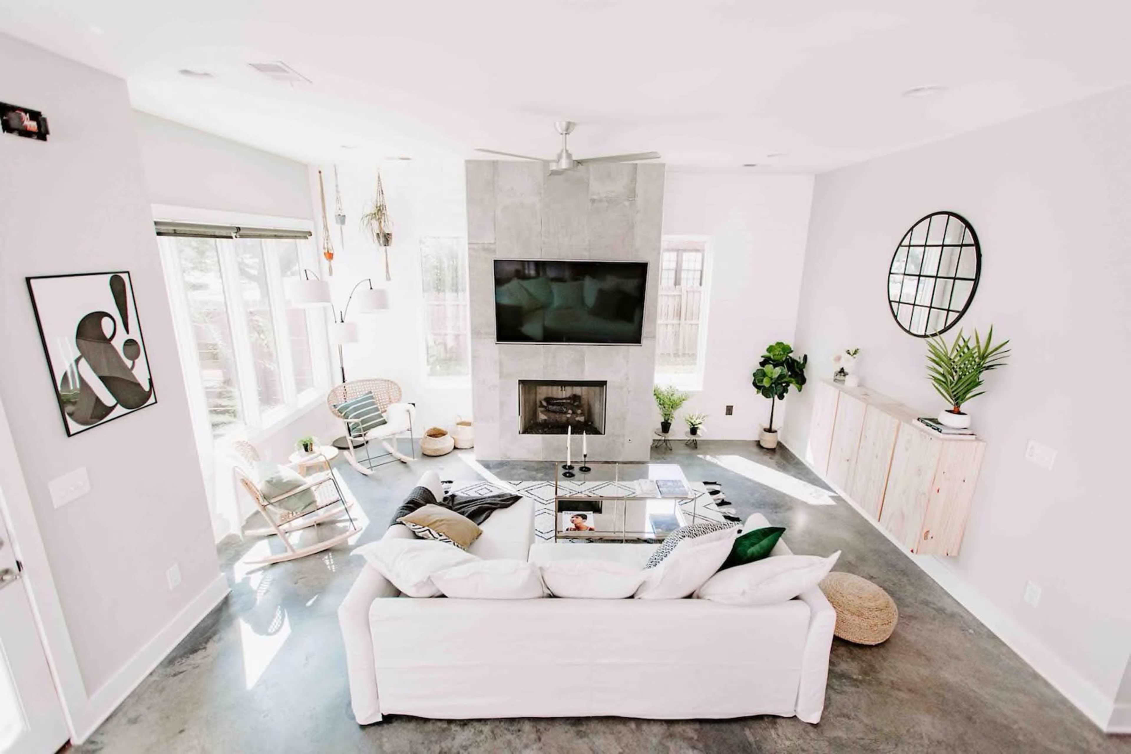 A bright living room features a white sofa facing a modern fireplace with a mounted television above, surrounded by large windows and minimalist decor.