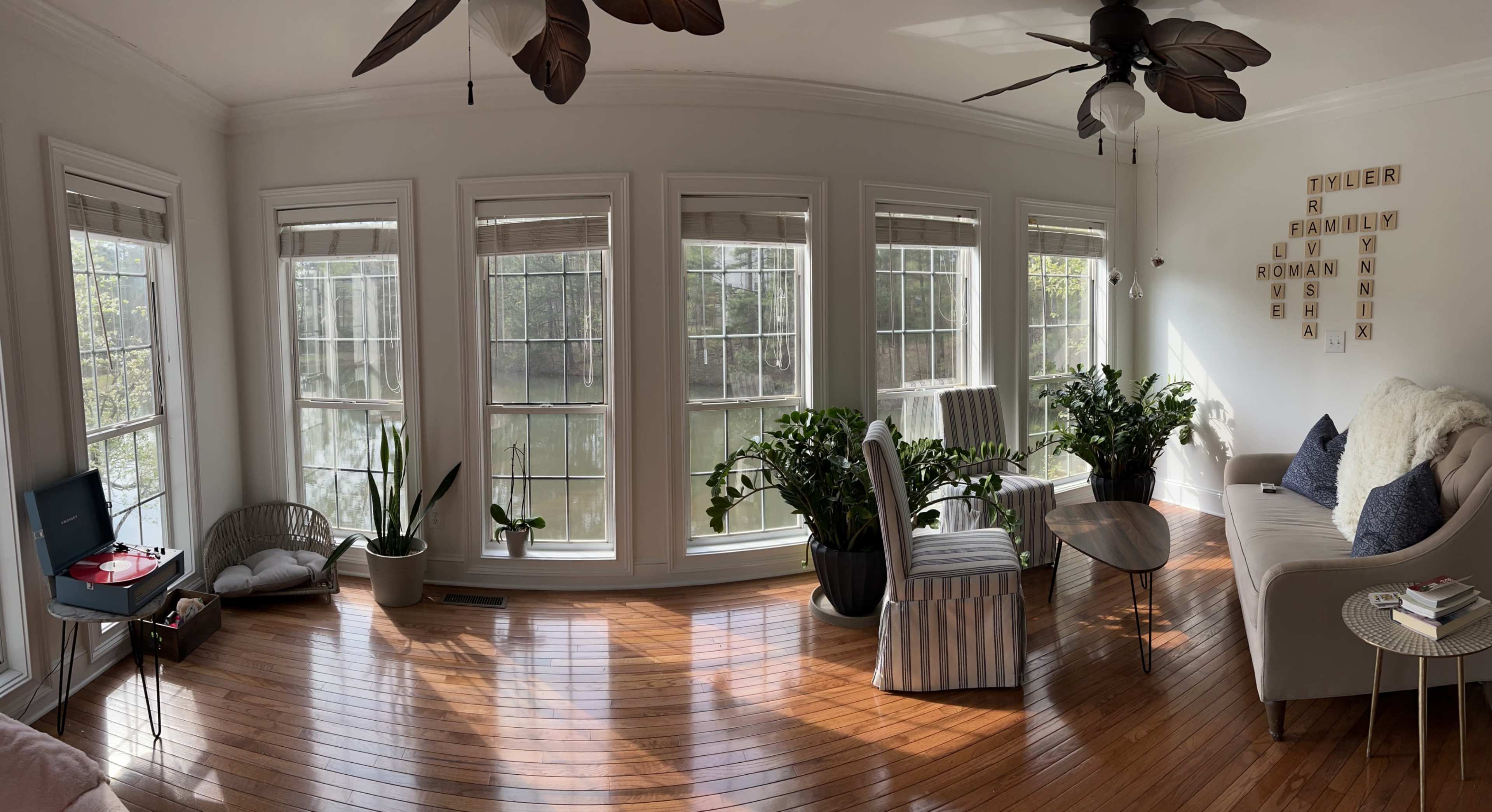 The room features hardwood floors and large windows overlooking a body of water, with plants, a record player, and a striped chair arranged around a small table.