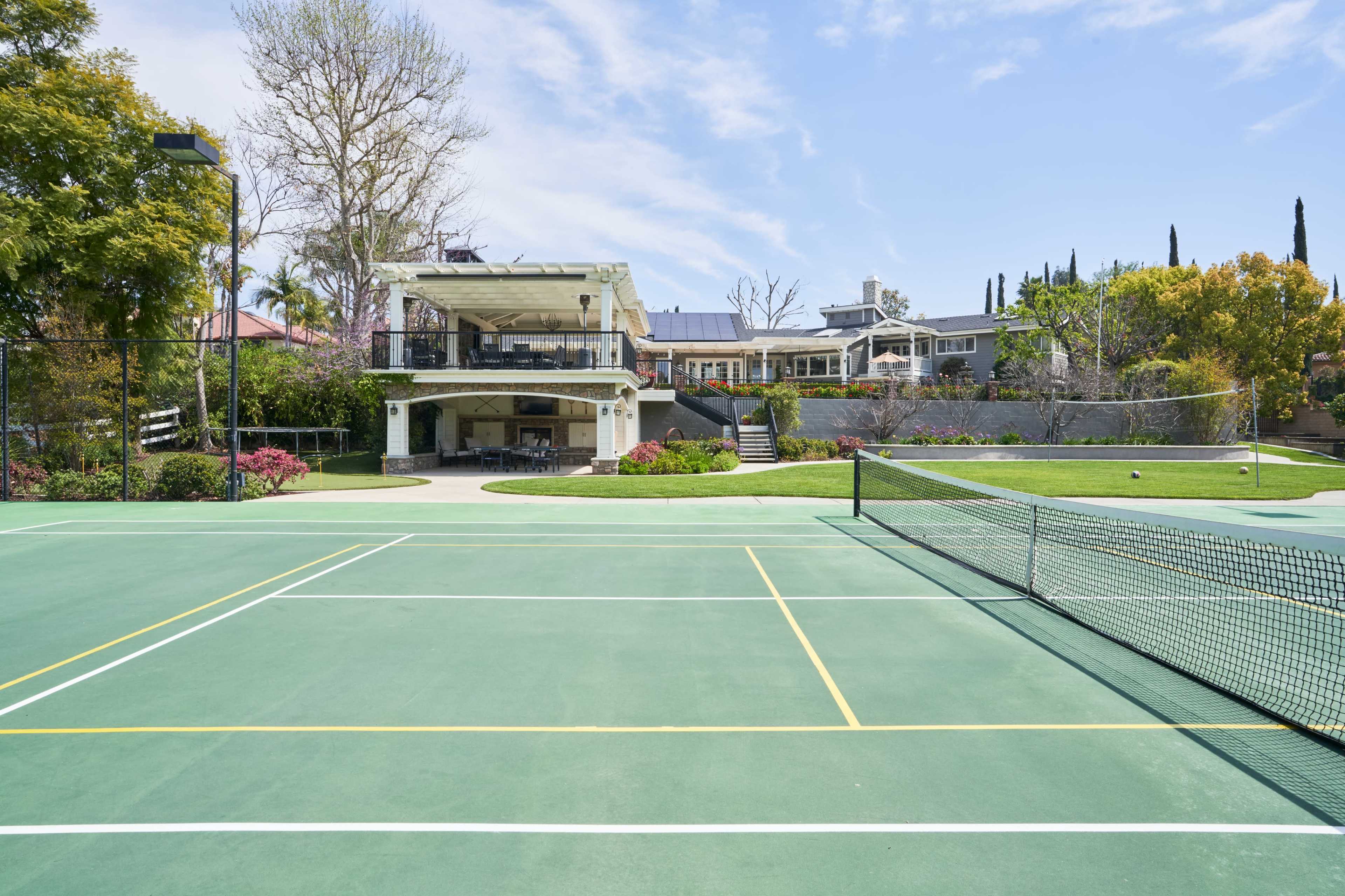 The image shows a well-maintained tennis court in the foreground with a large, modern home and landscaped garden in the background.