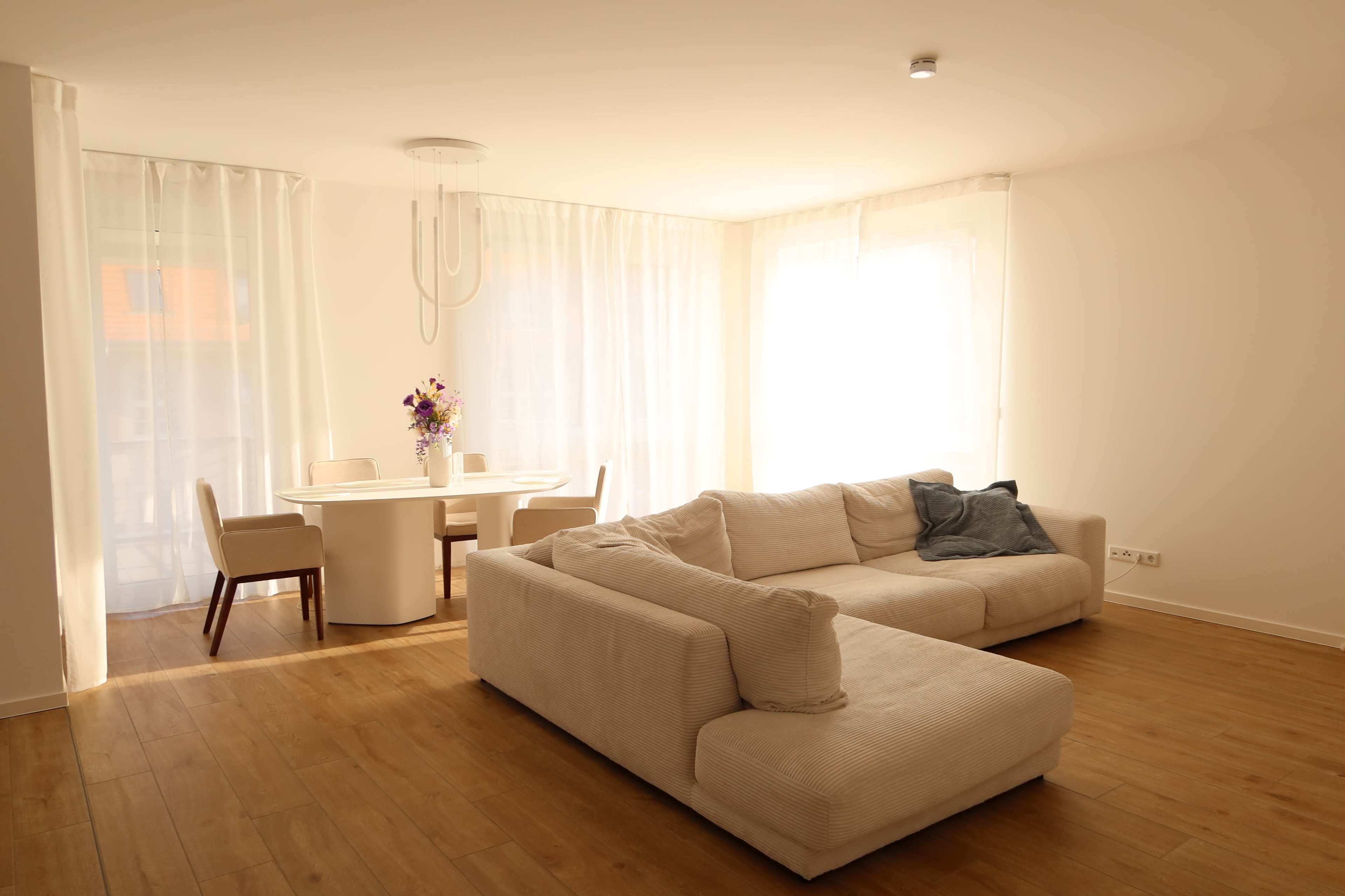 The image shows a bright, minimalist living space with a large beige sofa, a round dining table, and white curtains letting in natural light.