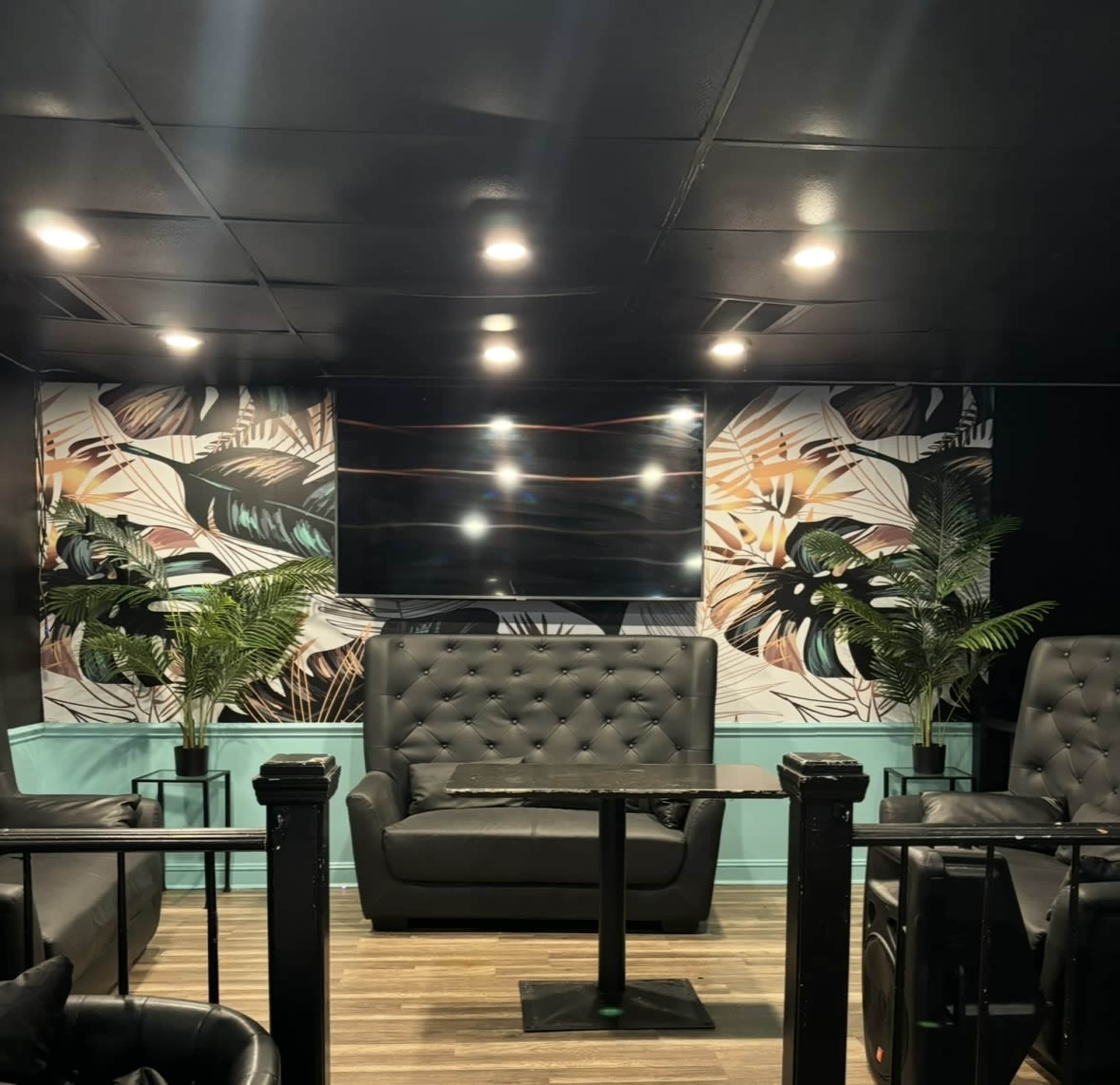 The image shows a dimly lit lounge area with black seating, a table, and tropical-themed wall decor featuring plants.