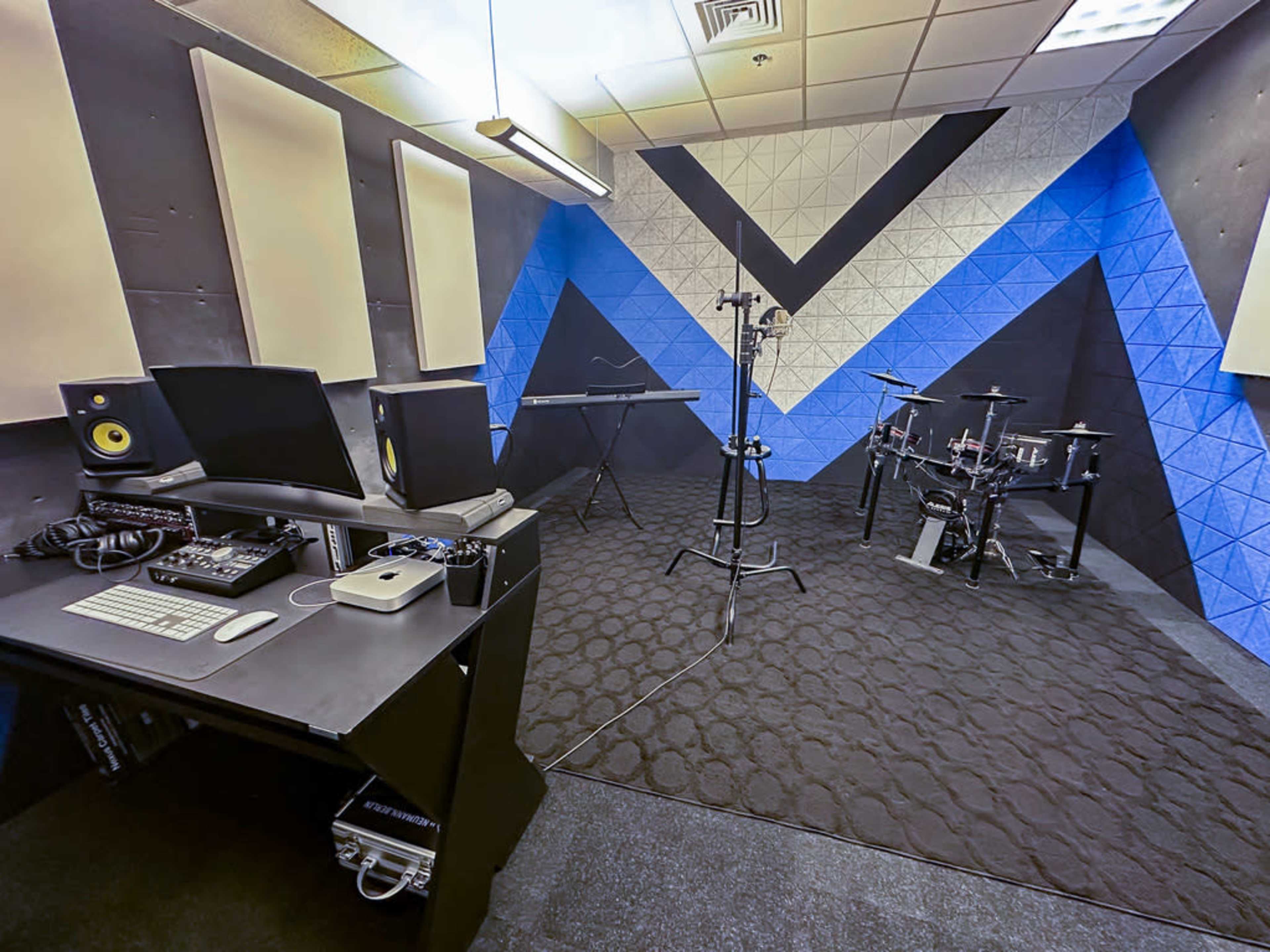 A music recording studio features a computer setup, a keyboard, and electronic drums against a patterned blue and black wall.