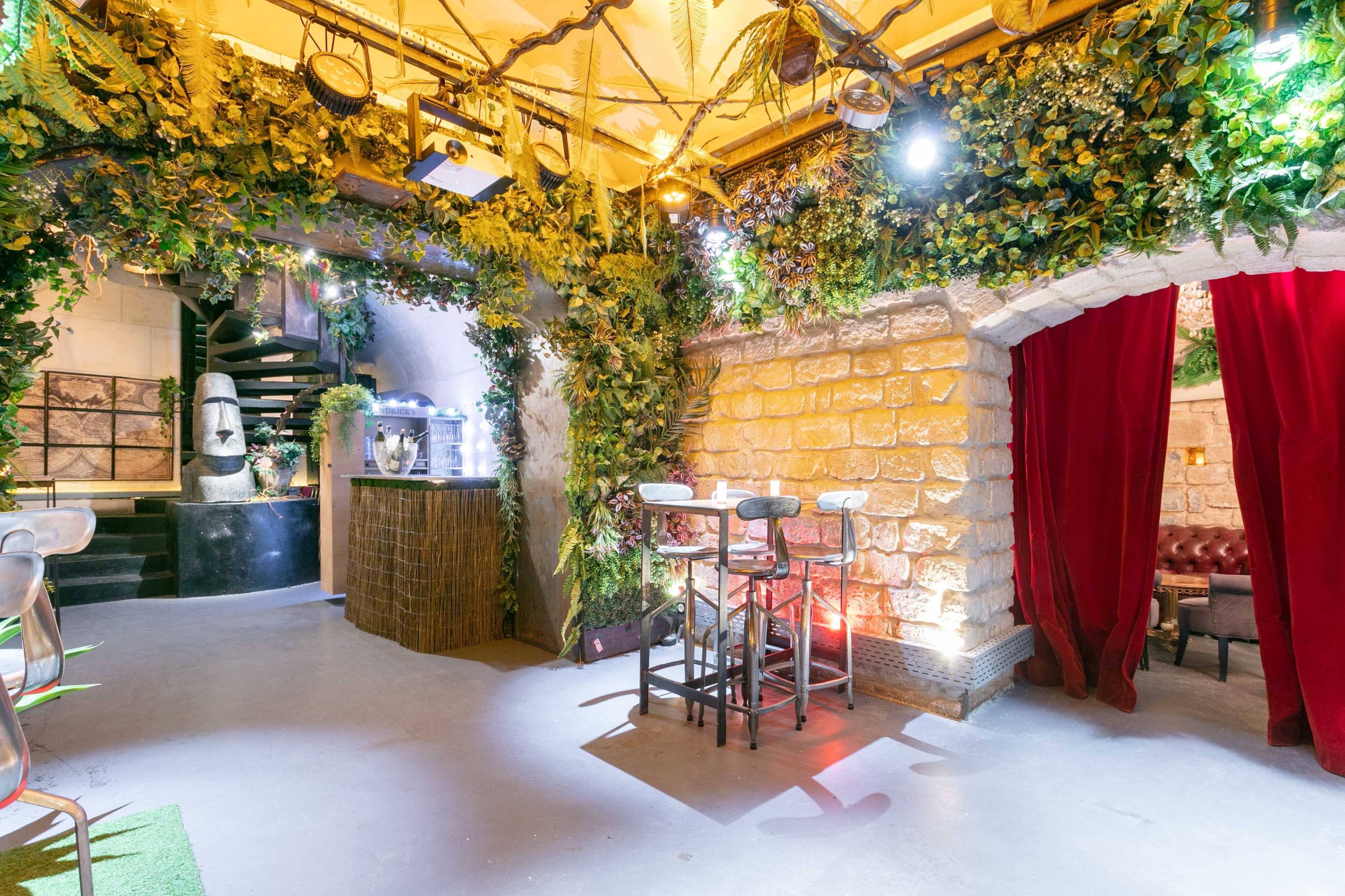 A dimly lit bar area featuring stone walls, lush greenery, and a small seating arrangement under soft lighting.