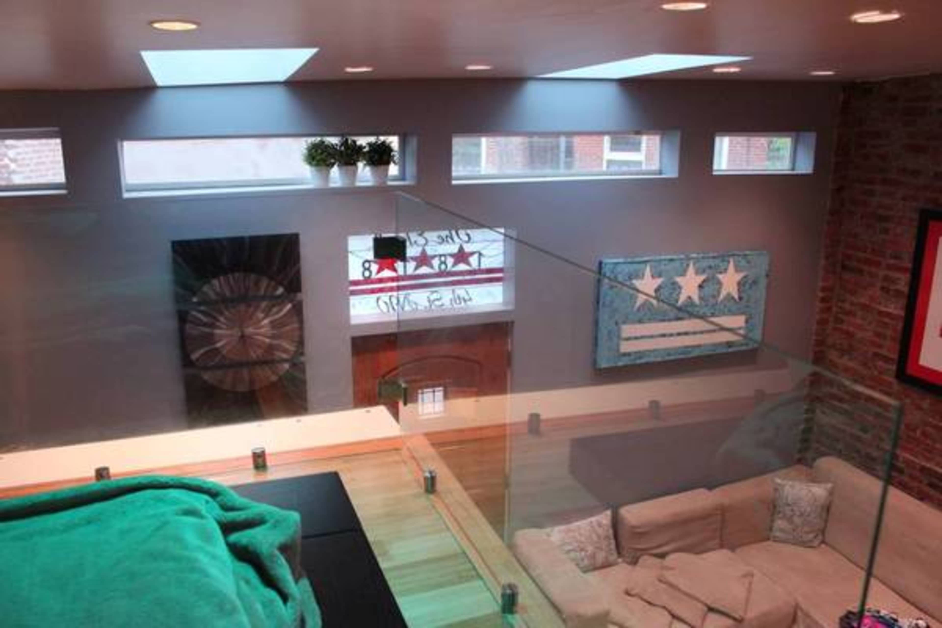 A modern living room featuring a glass railing, a couch, and decorative artwork on the walls, with natural light coming in from small windows above.
