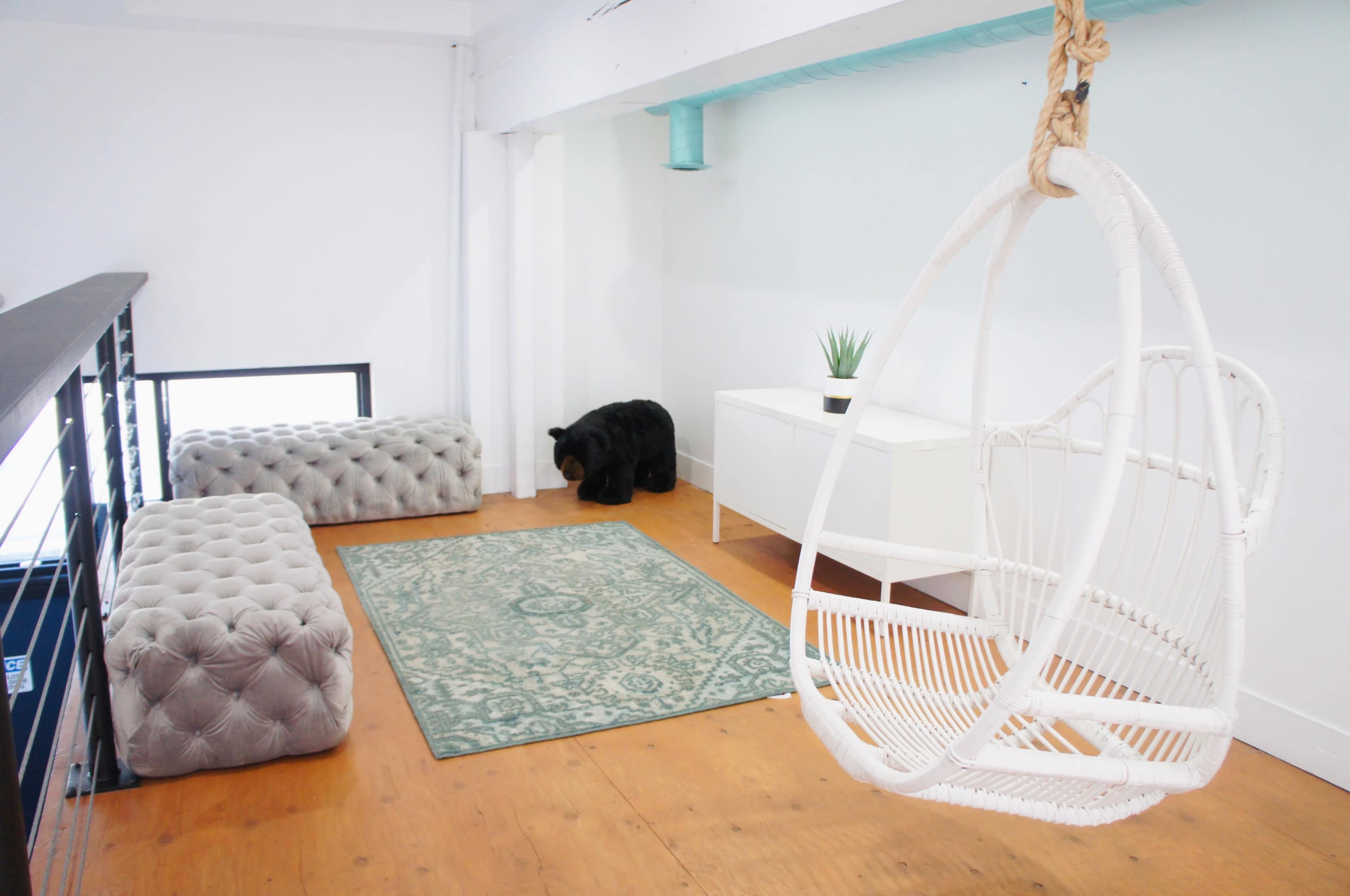 The image shows a modern indoor space featuring a hanging chair, a plush tufted ottoman, a small bear figurine, and a decorative rug on wooden flooring.