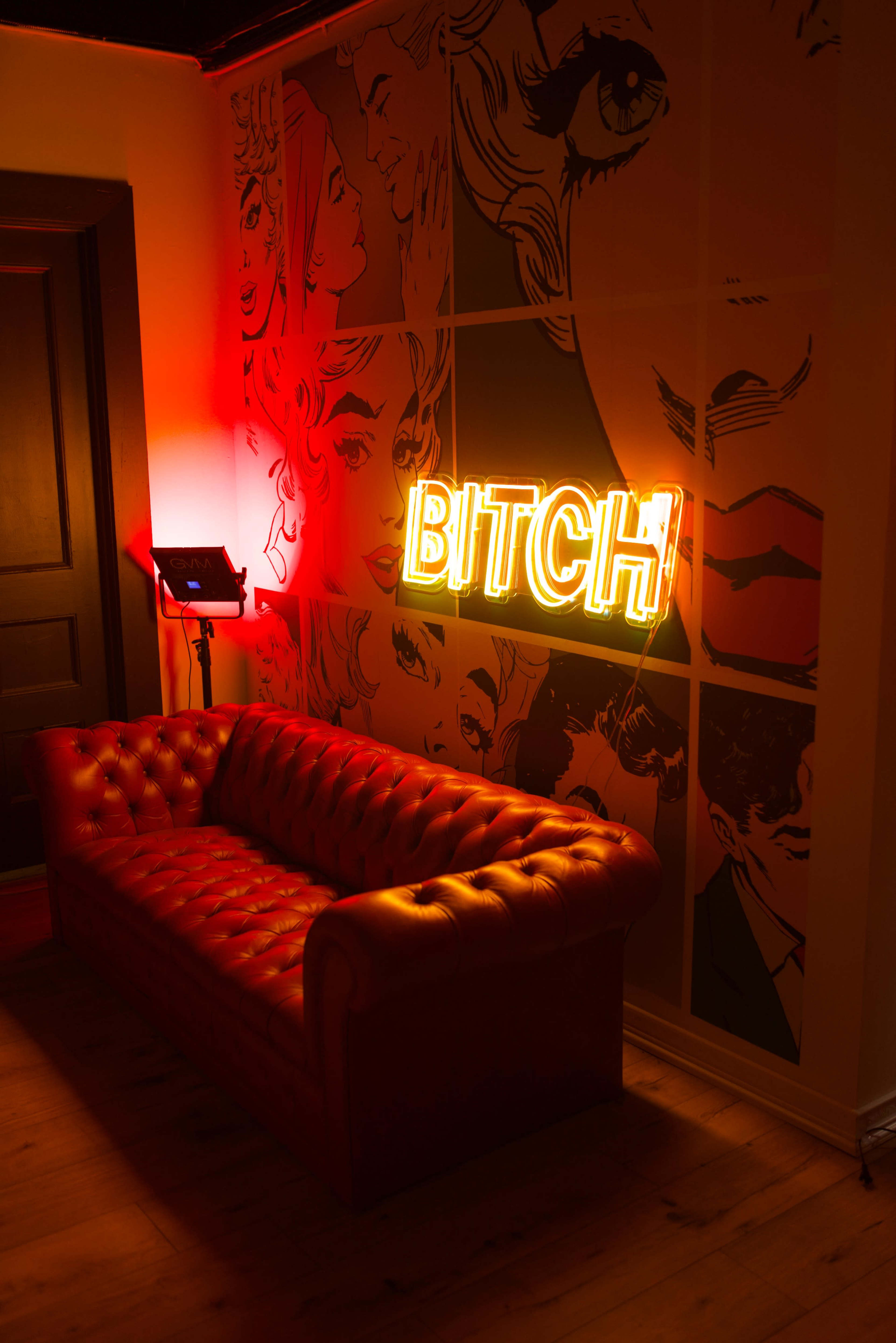A neon sign that reads "BITCH" is illuminated on a wall featuring graphic portraits, next to a red leather chesterfield sofa.