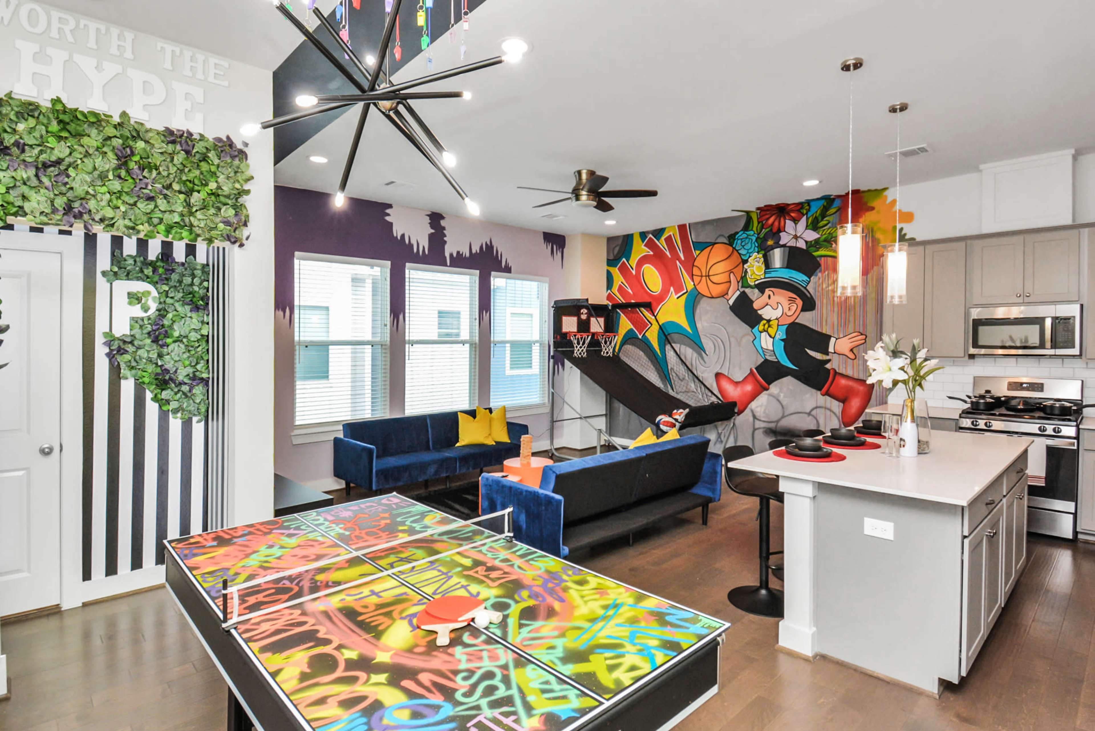 A brightly decorated modern living space featuring a kitchen, a lounge area with blue sofas, and a colorful mural on the wall.
