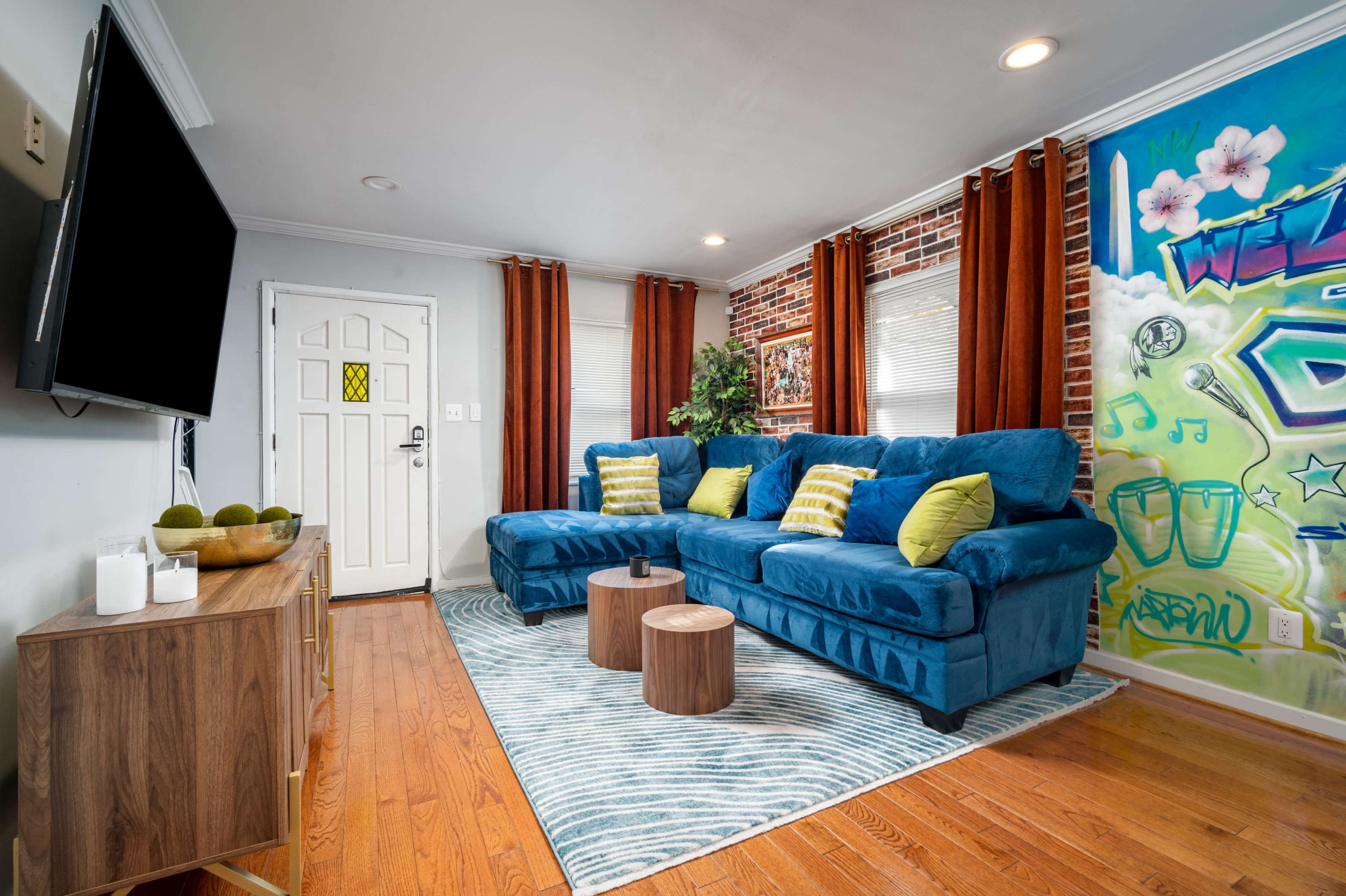 A blue sofa with yellow pillows is positioned in a living room featuring a brick wall, an abstract mural, and wooden furniture.