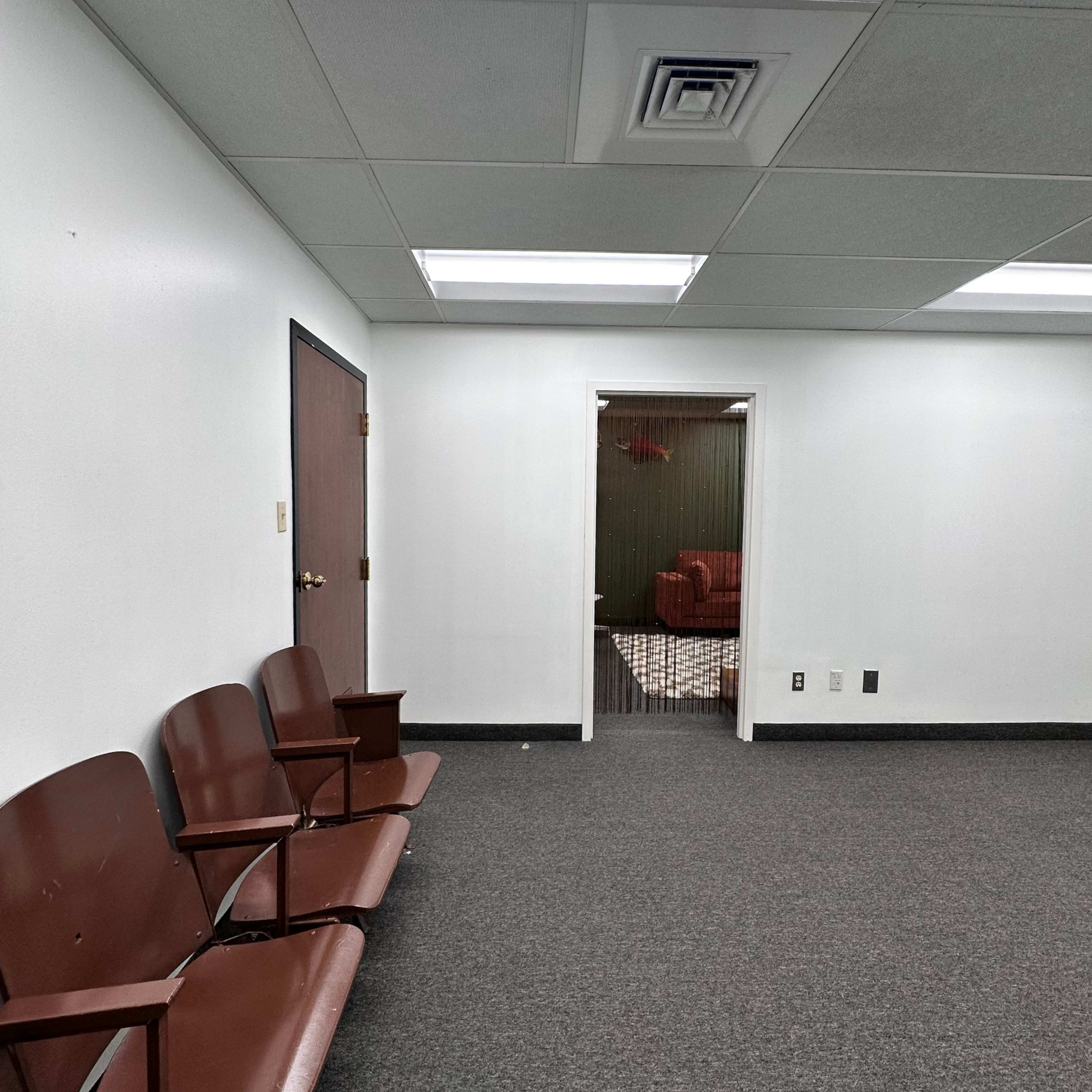The image shows an empty waiting room with brown chairs against a white wall and a doorway leading to another room in the background.