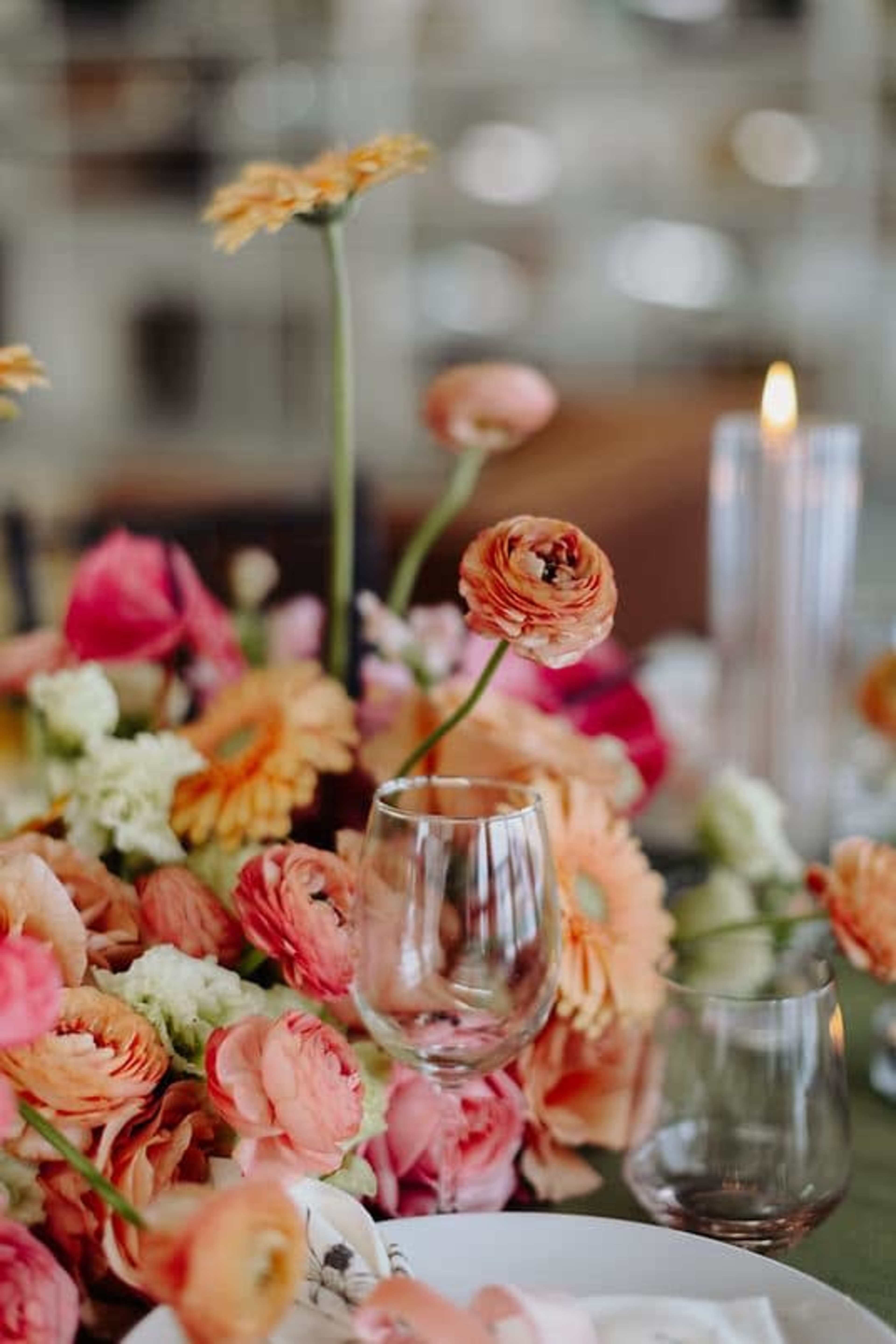 A vibrant floral centerpiece features a variety of colorful flowers, including ranunculus and gerbera daisies, arranged around elegant glassware and lit candles.