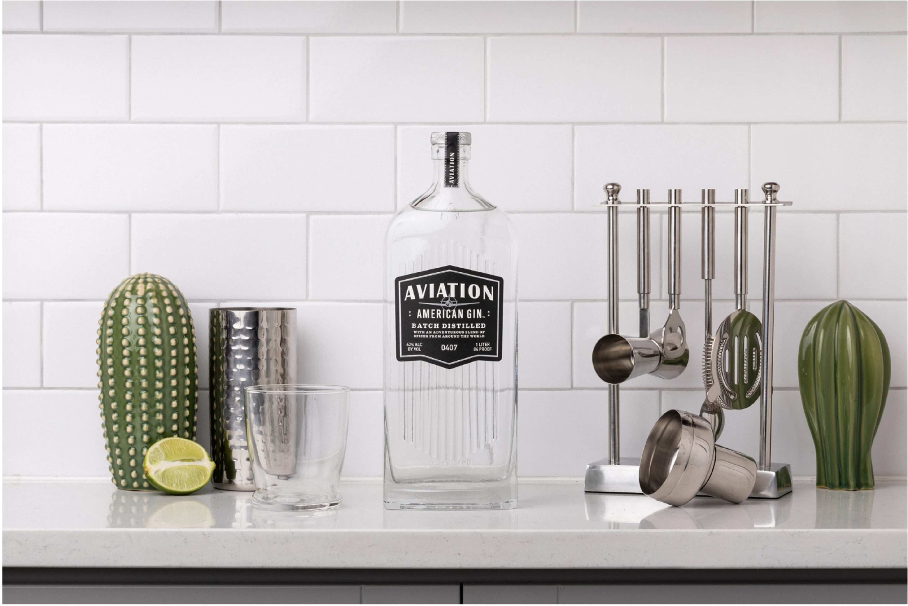 A bottle of Aviation American Gin stands on a countertop next to a glass, a lime half, a jigger, a strainer, and two decorative cacti.