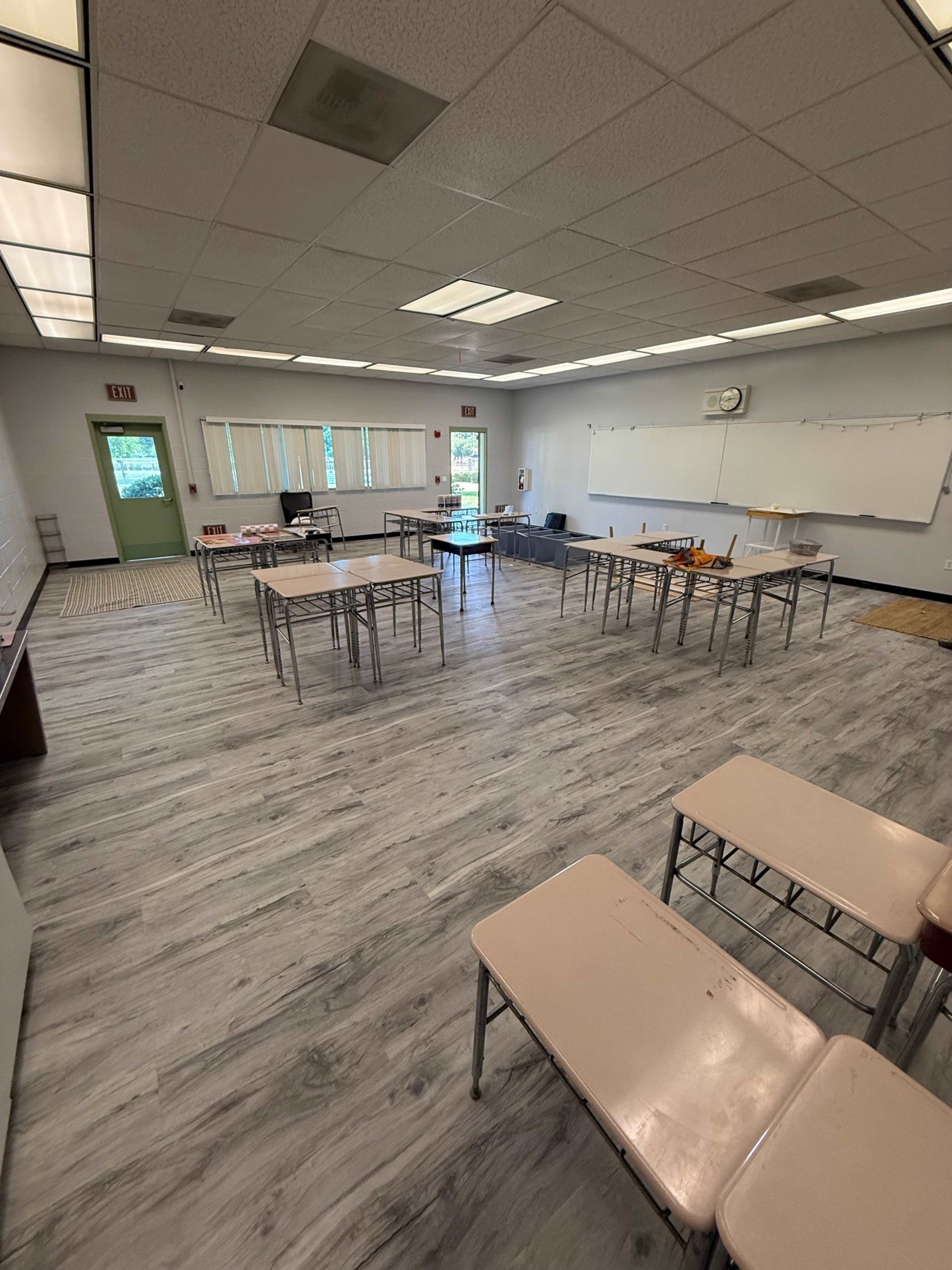 The image shows a classroom with several metal-framed tables arranged in a scattered layout on a wooden-looking floor.