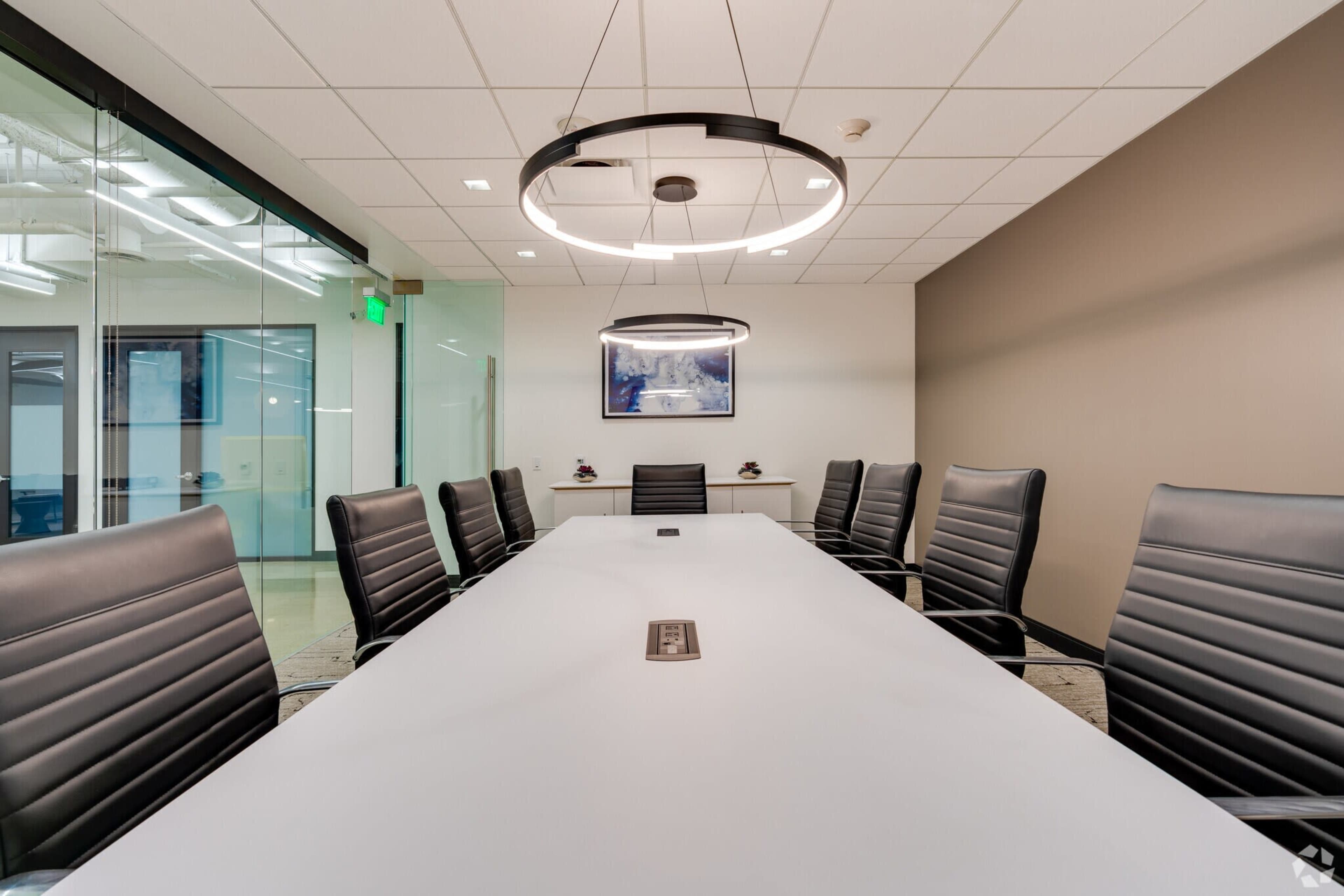 A long conference table with black leather chairs is positioned in a modern meeting room featuring overhead light fixtures and a wall-mounted artwork.