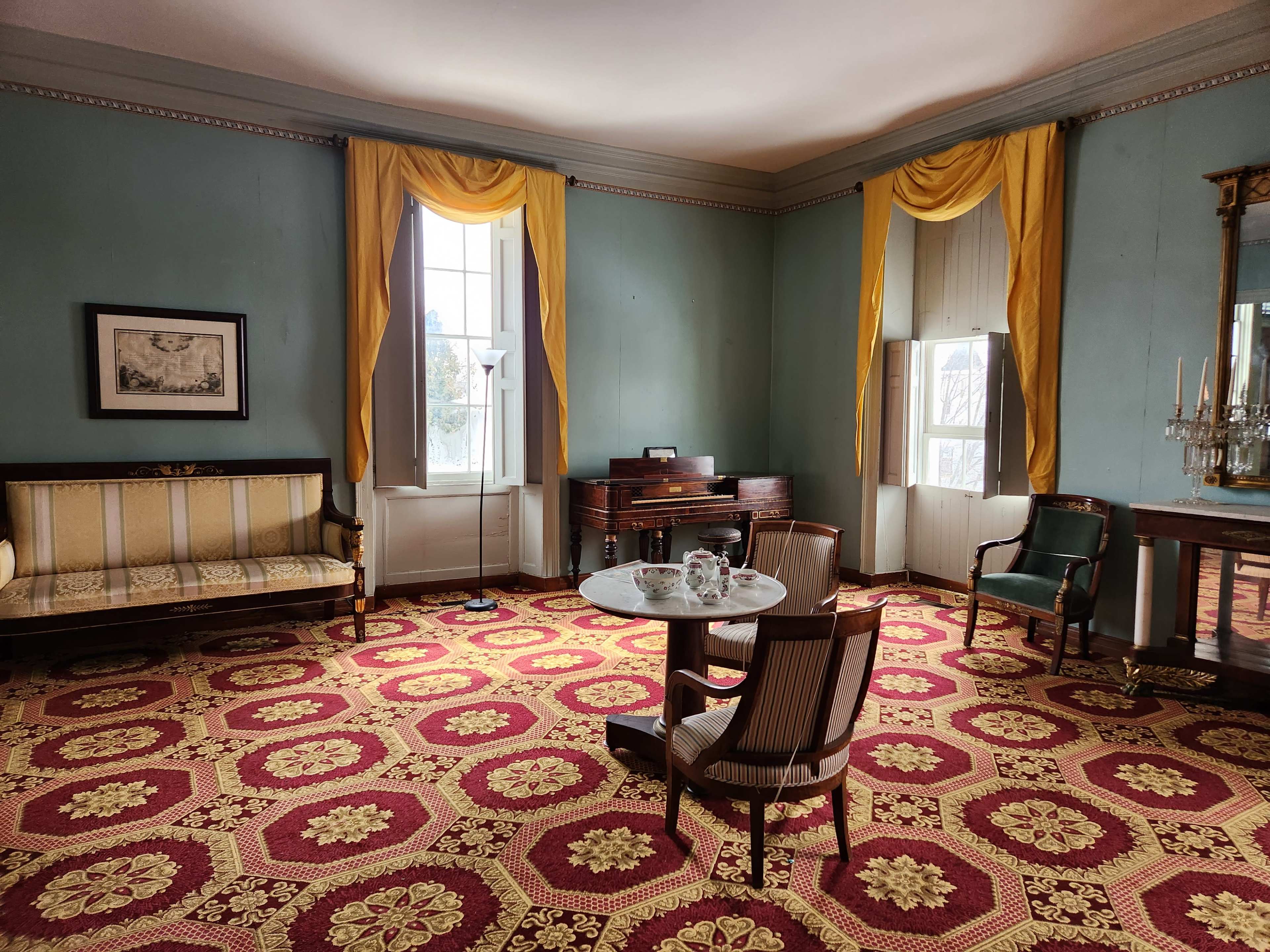 A vintage room with green walls, yellow drapes, a patterned carpet, a round table, and a sofa along with a piano in the background.