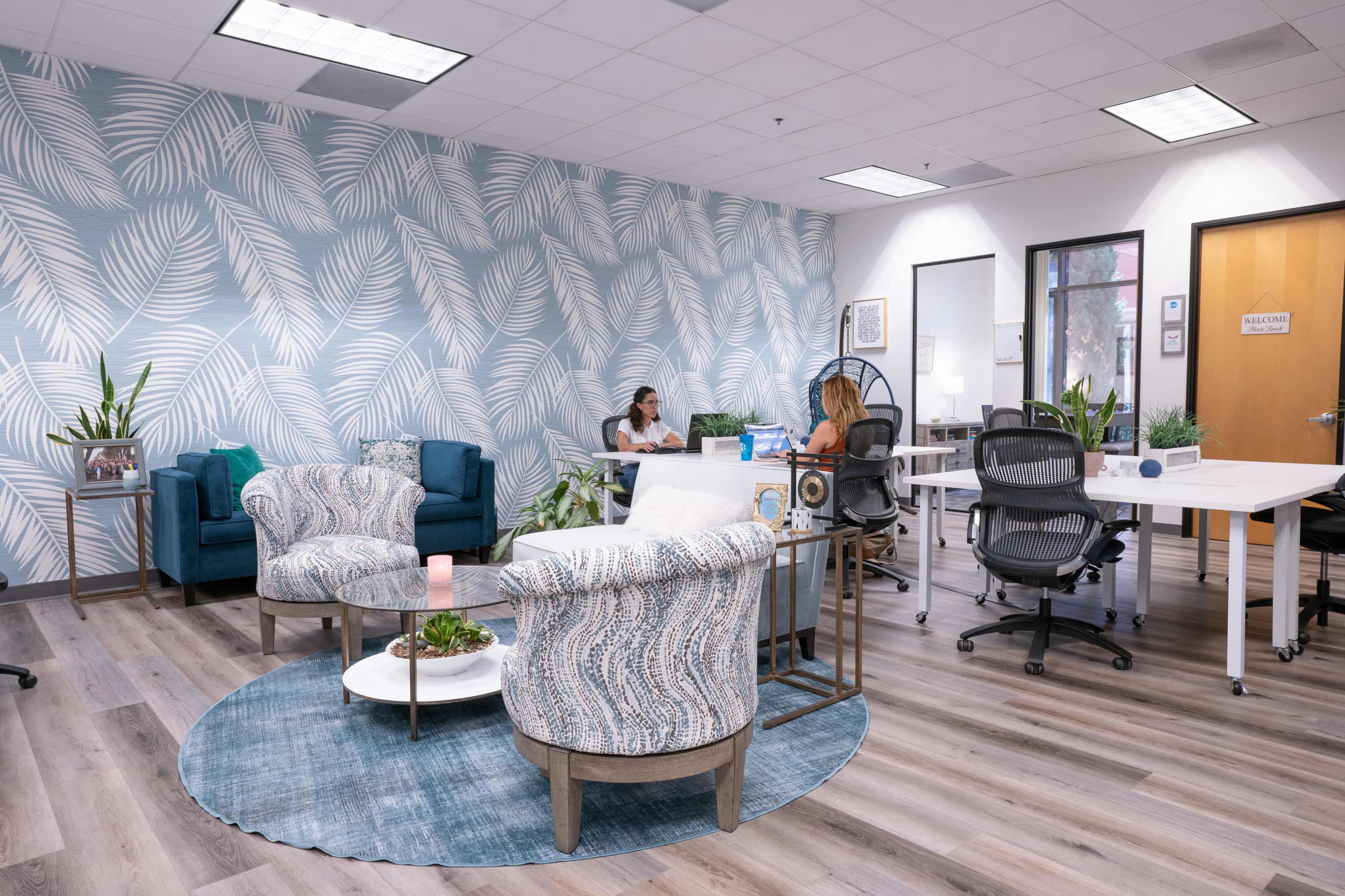 The image shows a modern office space featuring blue and white patterned wallpaper, a circular rug, and various seating arrangements including chairs and desks, with two women engaged in conversation.