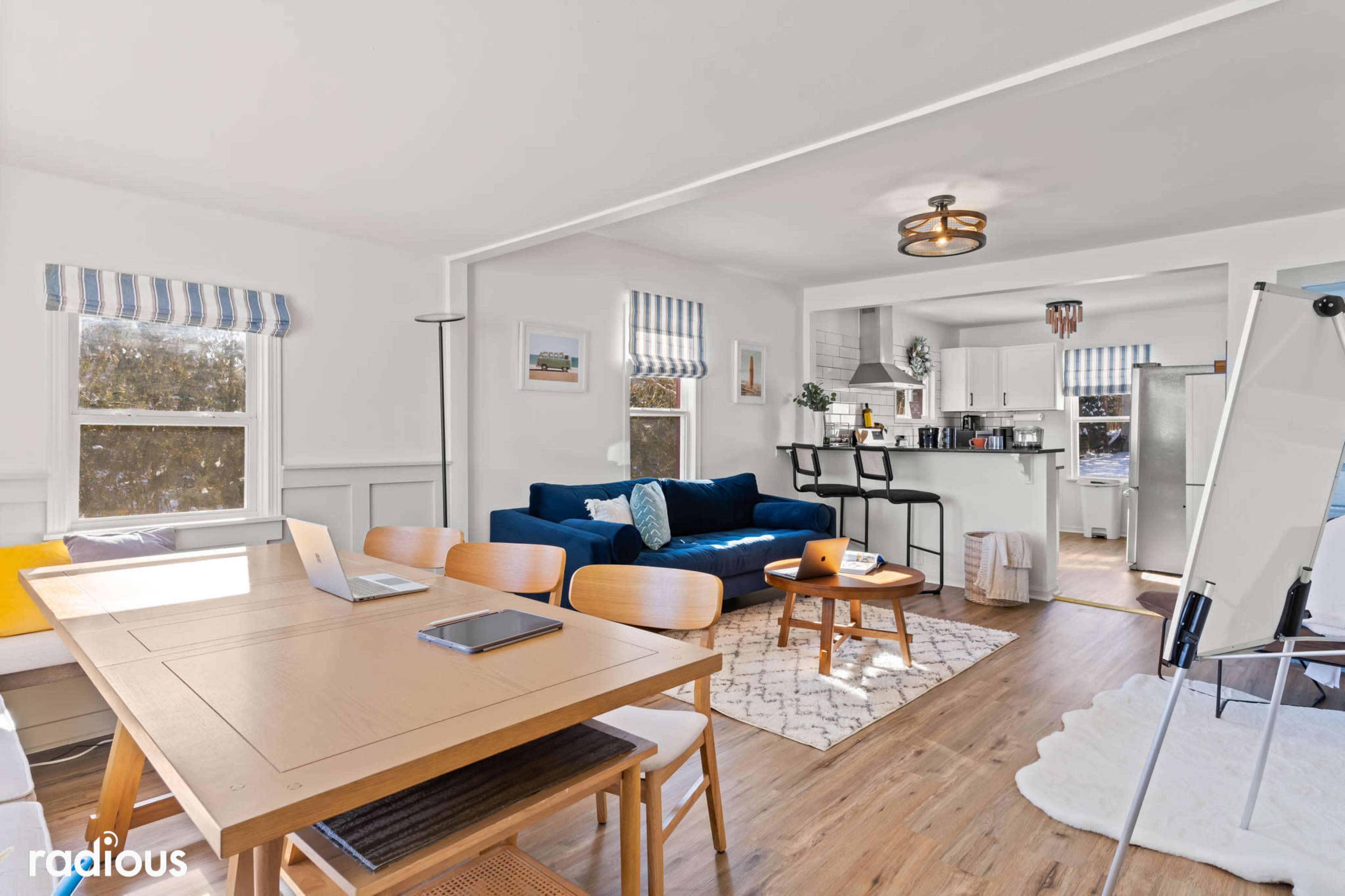 A bright and airy living space features a blue sofa, a dining table with chairs, and a kitchen area visible in the background.