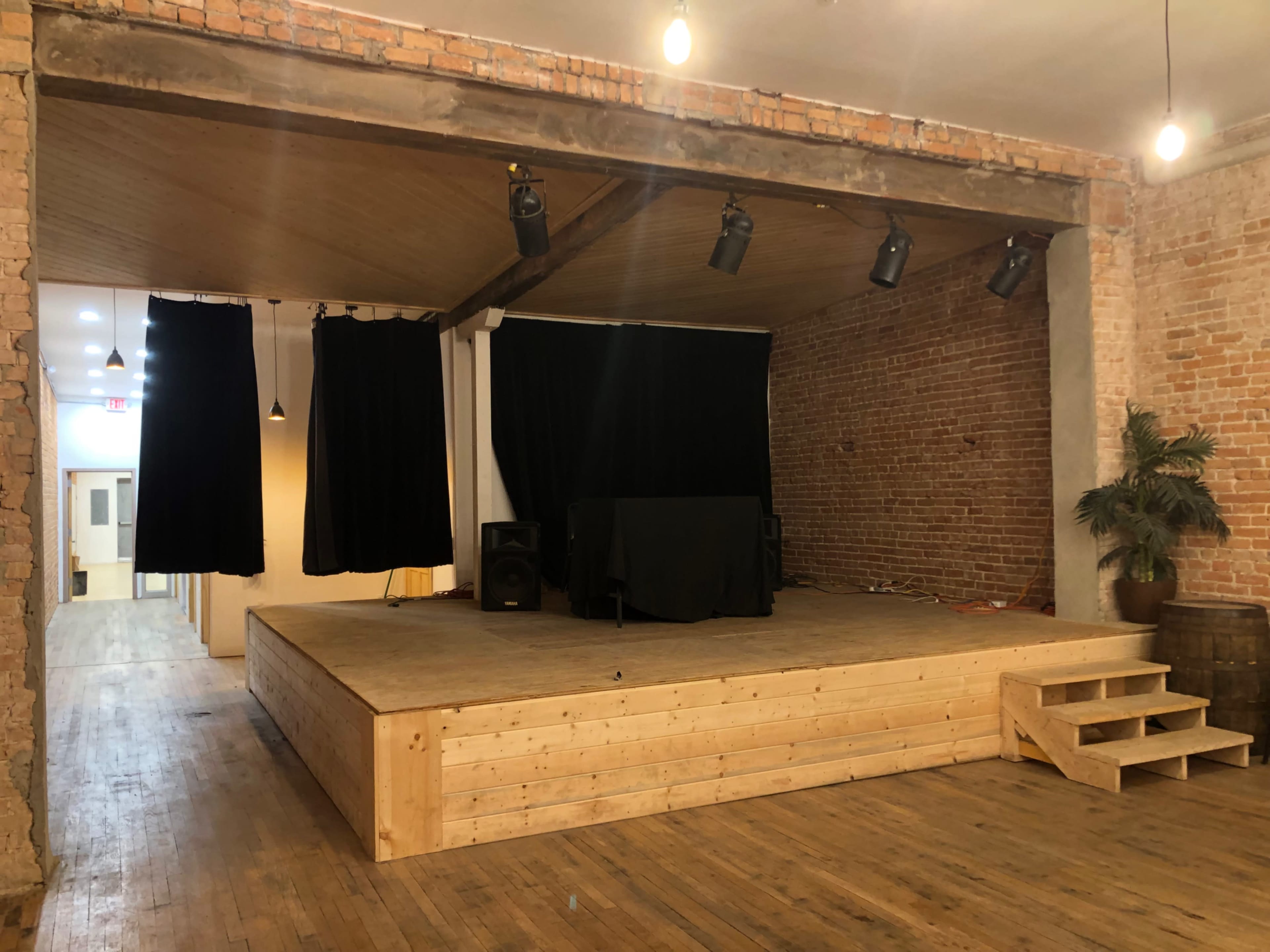 A wooden stage with a black backdrop and sound equipment, situated in a room with exposed brick walls and wooden flooring.