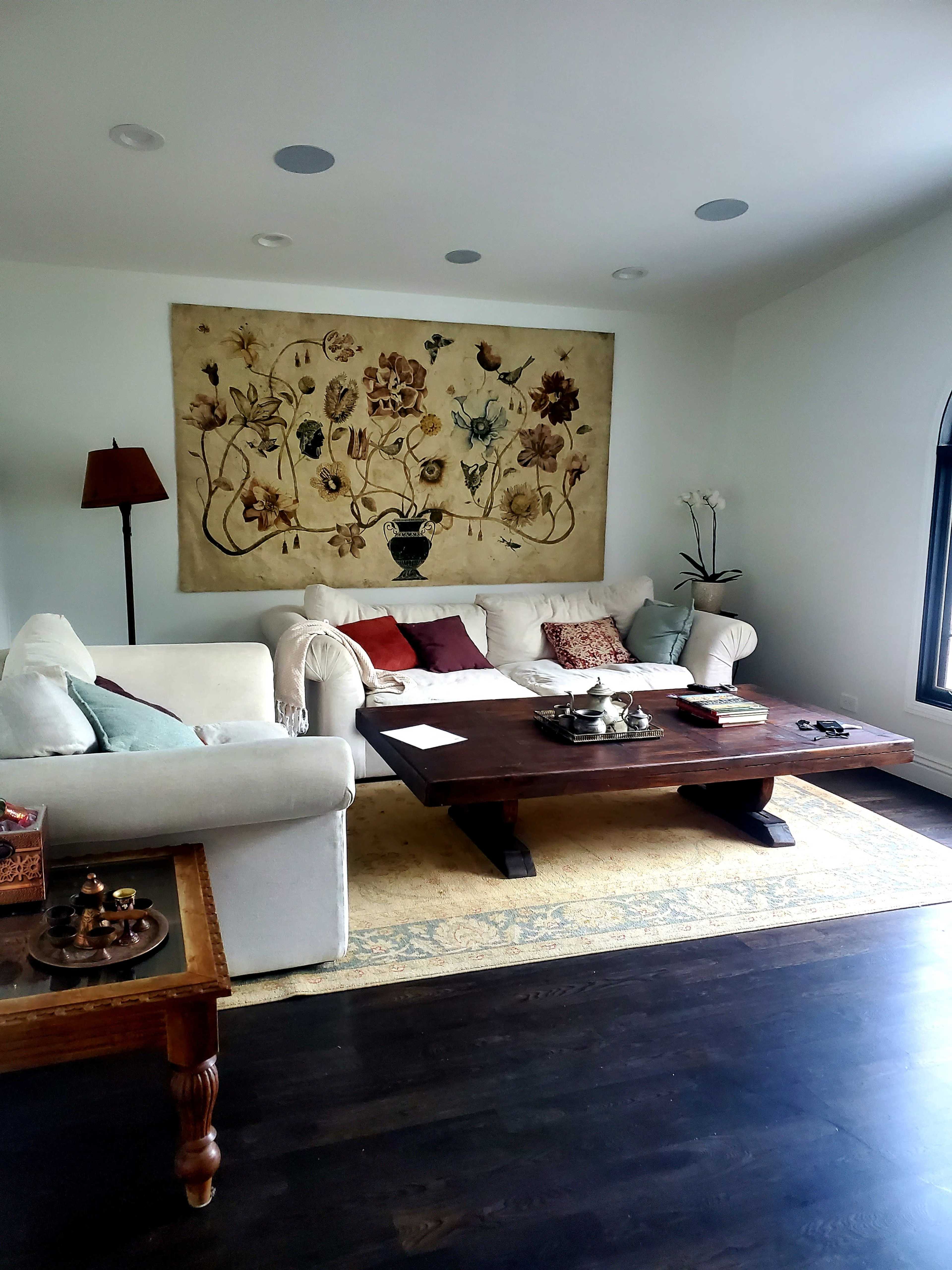 The image shows a living room with a white couch, a wooden coffee table, an ornate wall hanging, and a lamp in the corner.