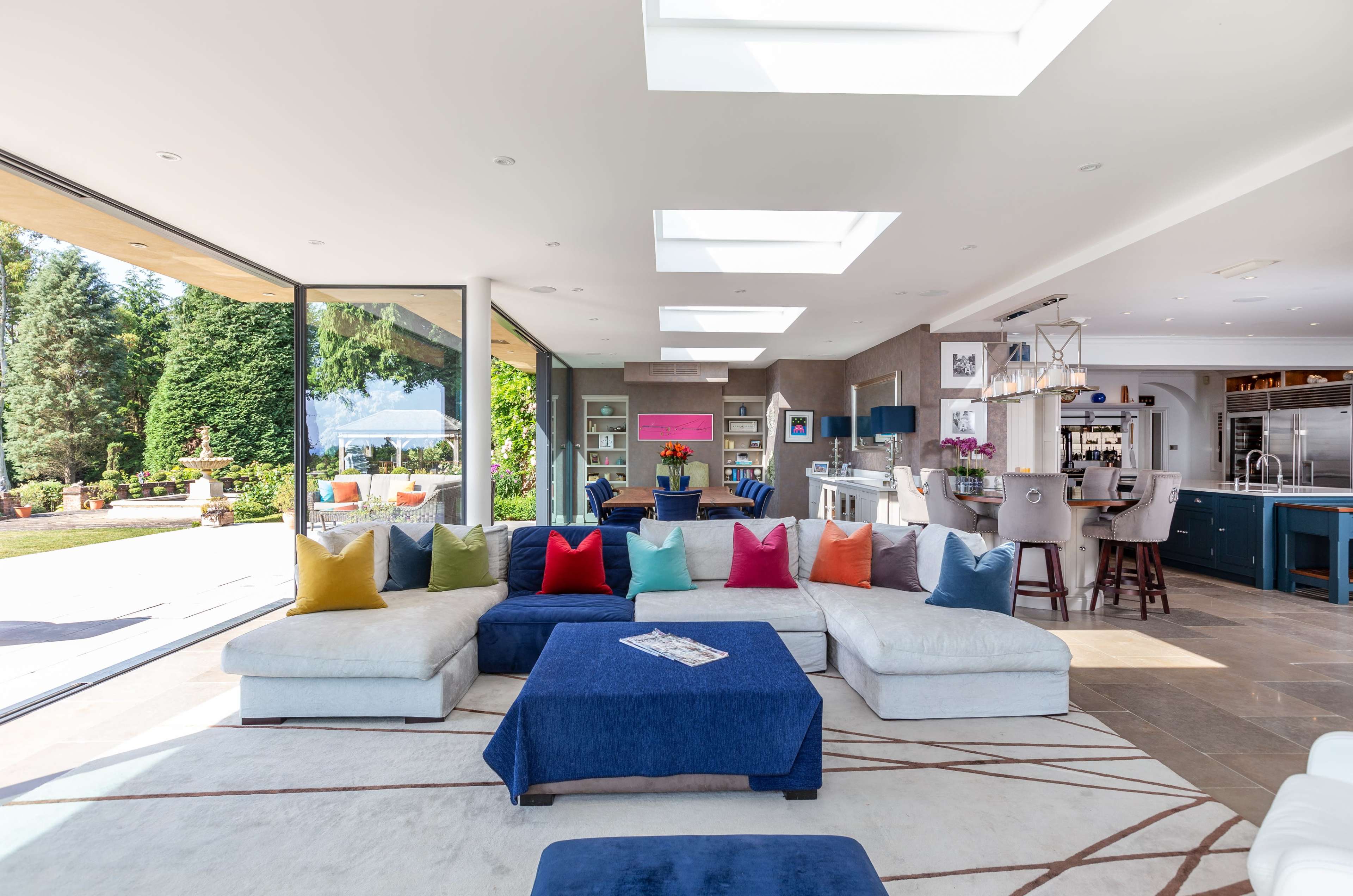 The image shows a spacious, modern living room with a large sectional sofa adorned with colorful cushions, overlooking a garden through expansive glass doors.