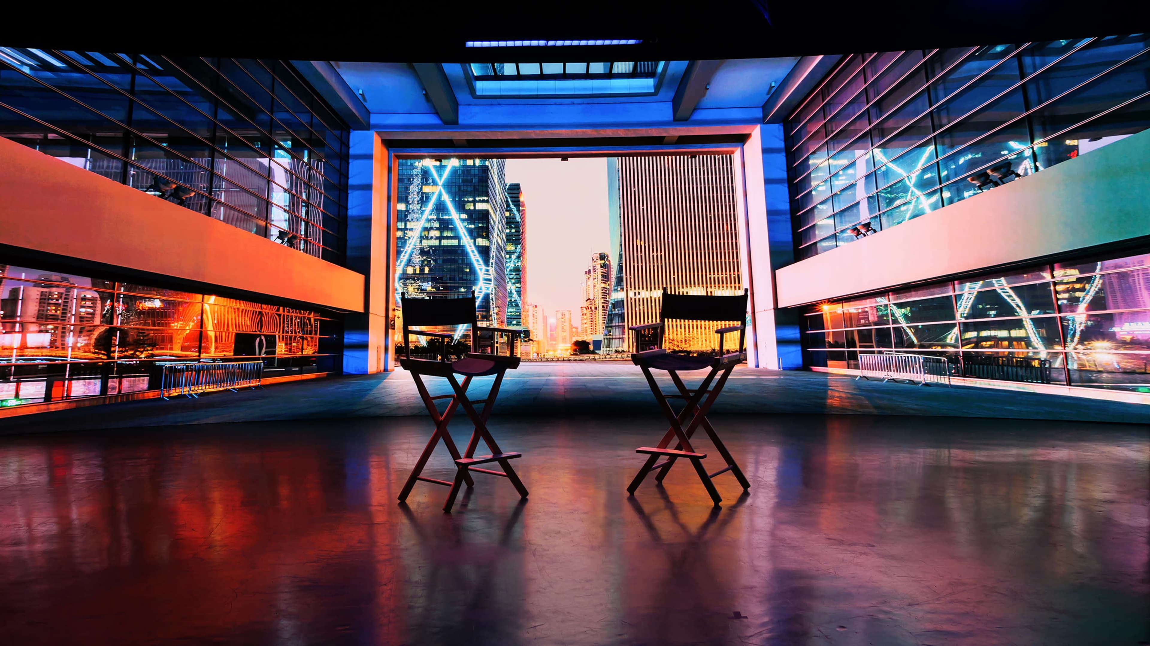 Two director's chairs are set facing a large window displaying a city skyline with brightly lit buildings at dusk.