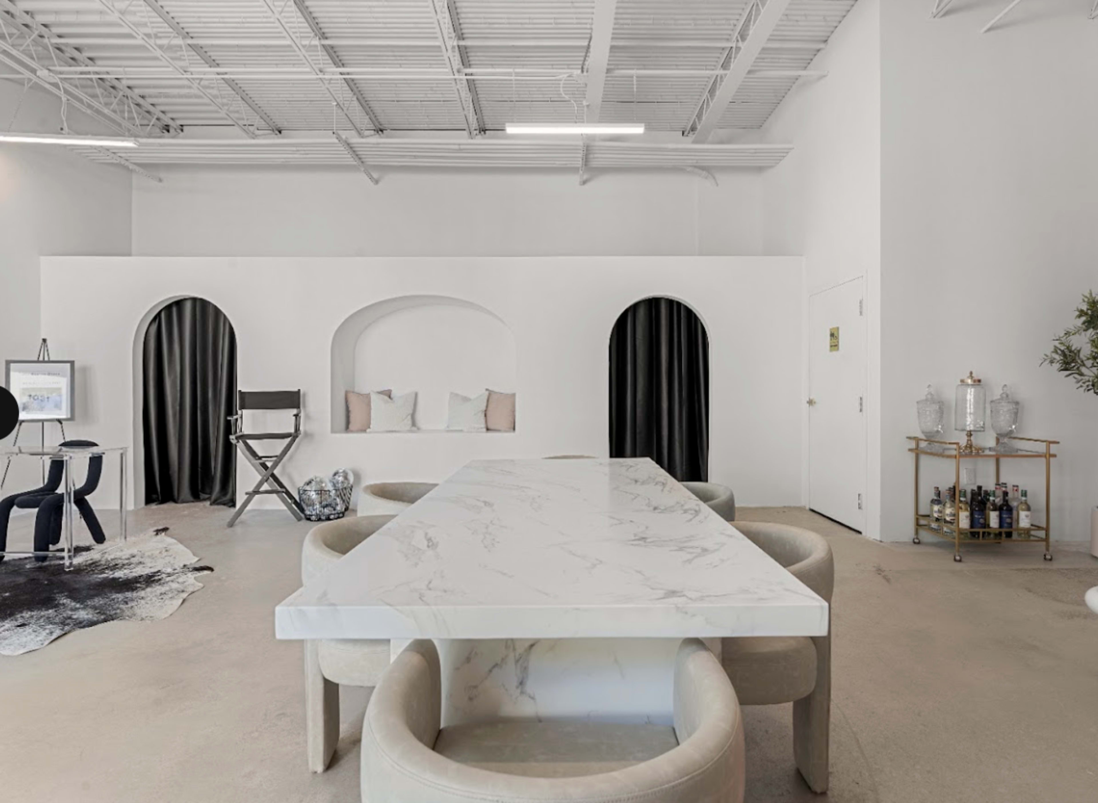 The scene features a spacious, minimalist room with a large marble dining table surrounded by light-colored upholstered chairs, flanked by arched niches and curtains.