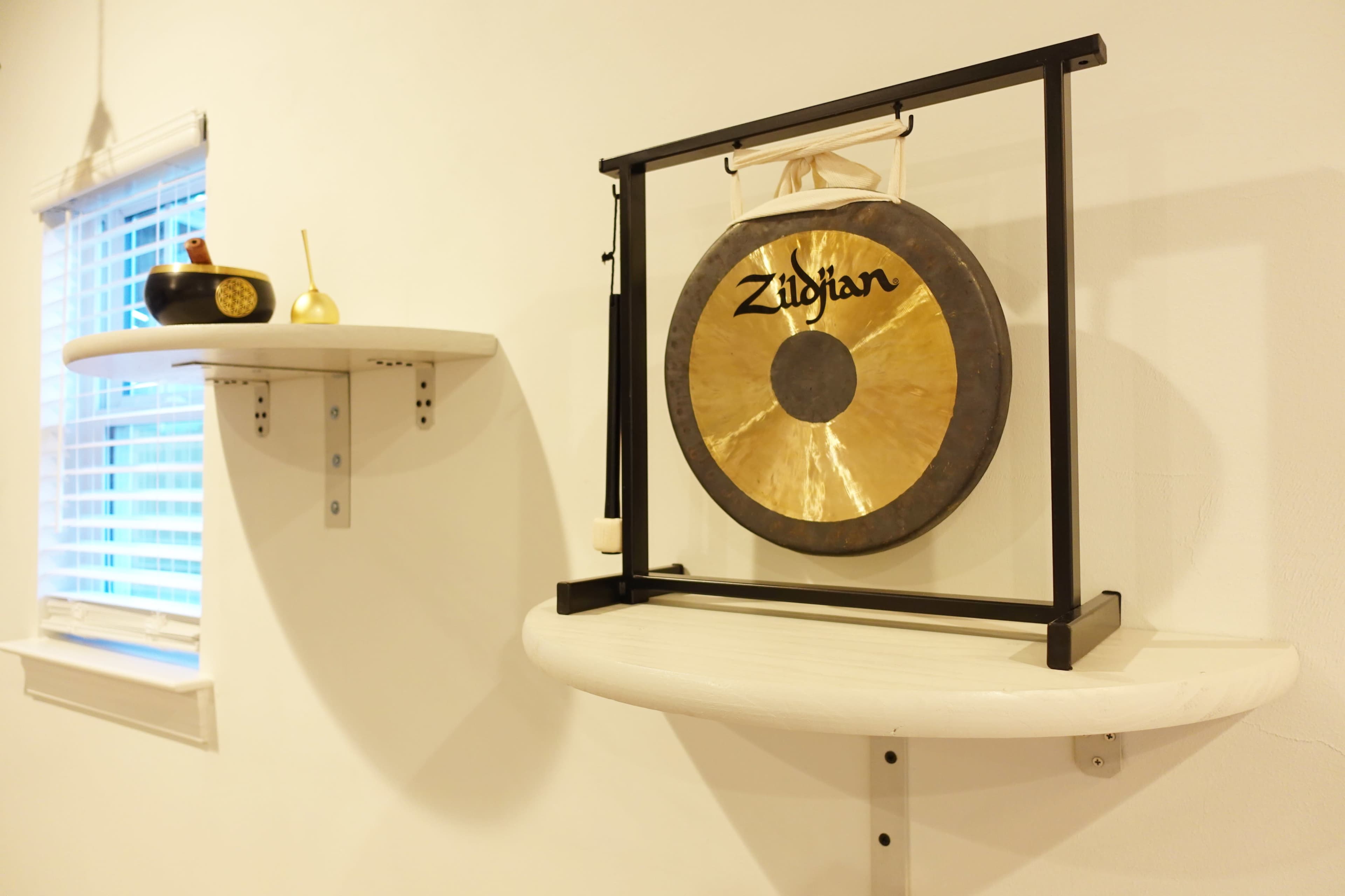 A Zildjian gong is mounted on a black metal stand between two wall shelves in a room with white walls.