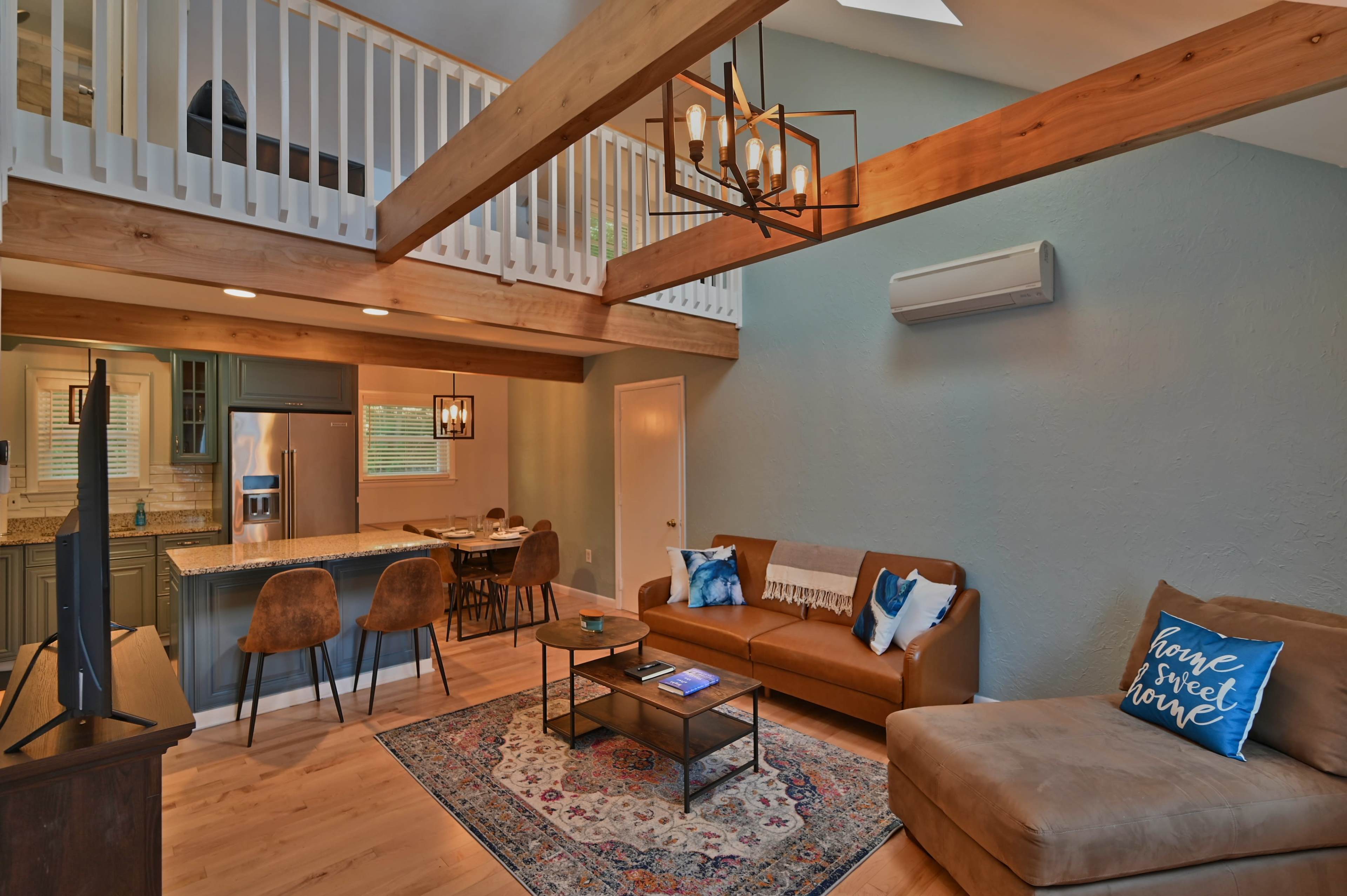The image shows a modern living room with a brown sofa, a coffee table, and an open kitchen area featuring bar stools and a dining set beneath wooden beams.
