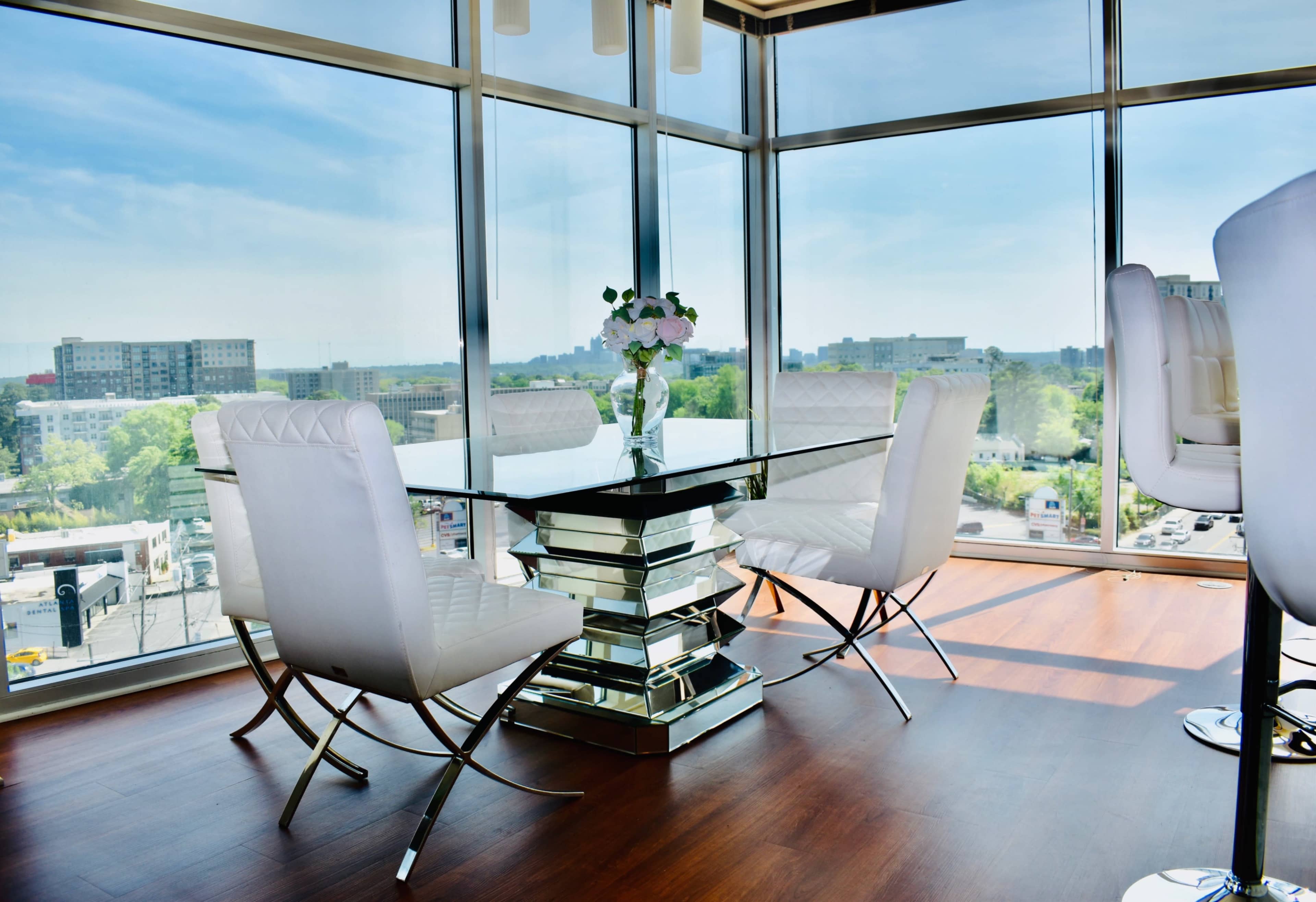 A modern dining area features a glass table surrounded by white chairs, situated in a bright space with large windows overlooking a cityscape.