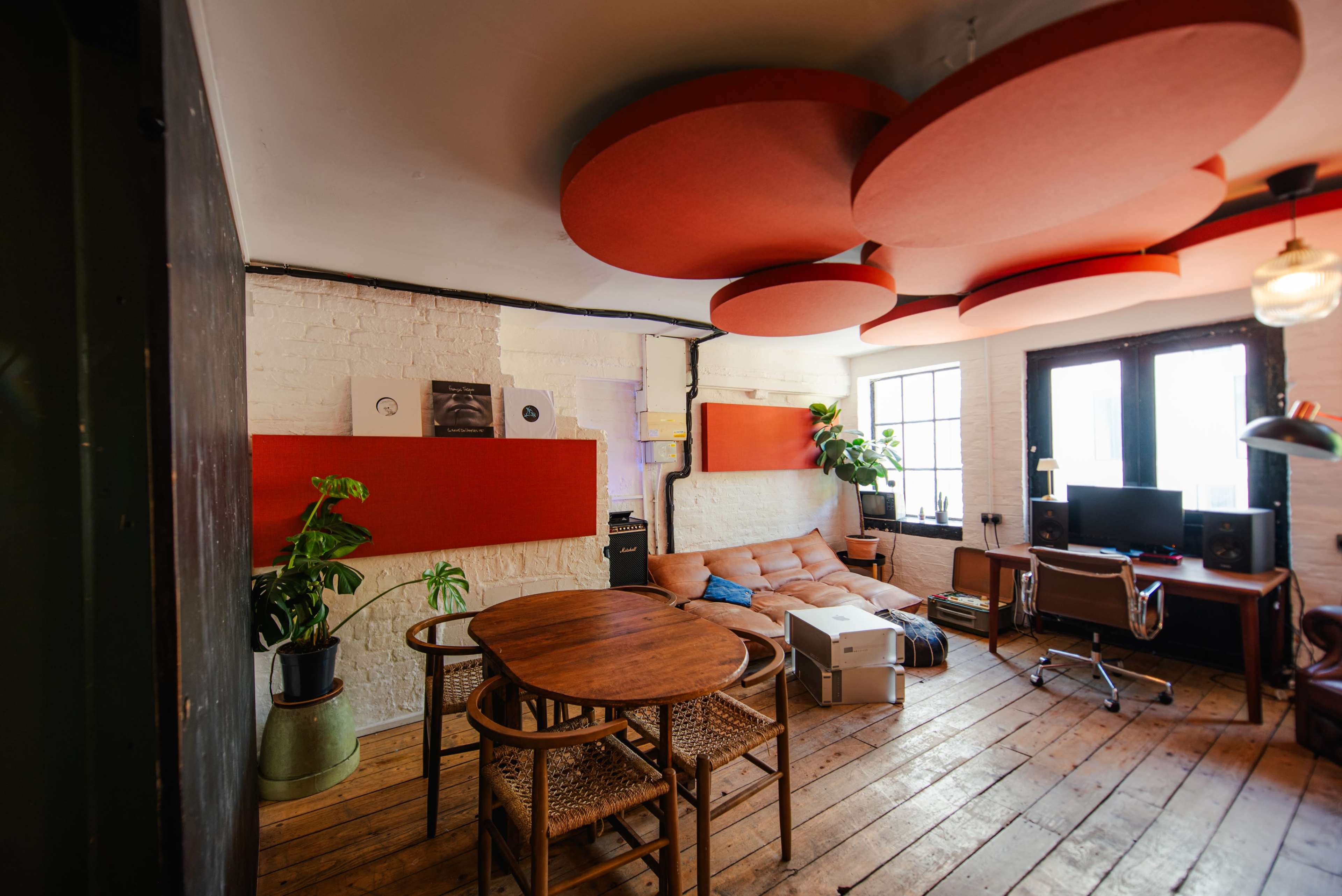 The image shows a cozy living room featuring a circular ceiling sound panel design, a light-colored sectional sofa, a wooden dining table, and a workspace with a computer setup.