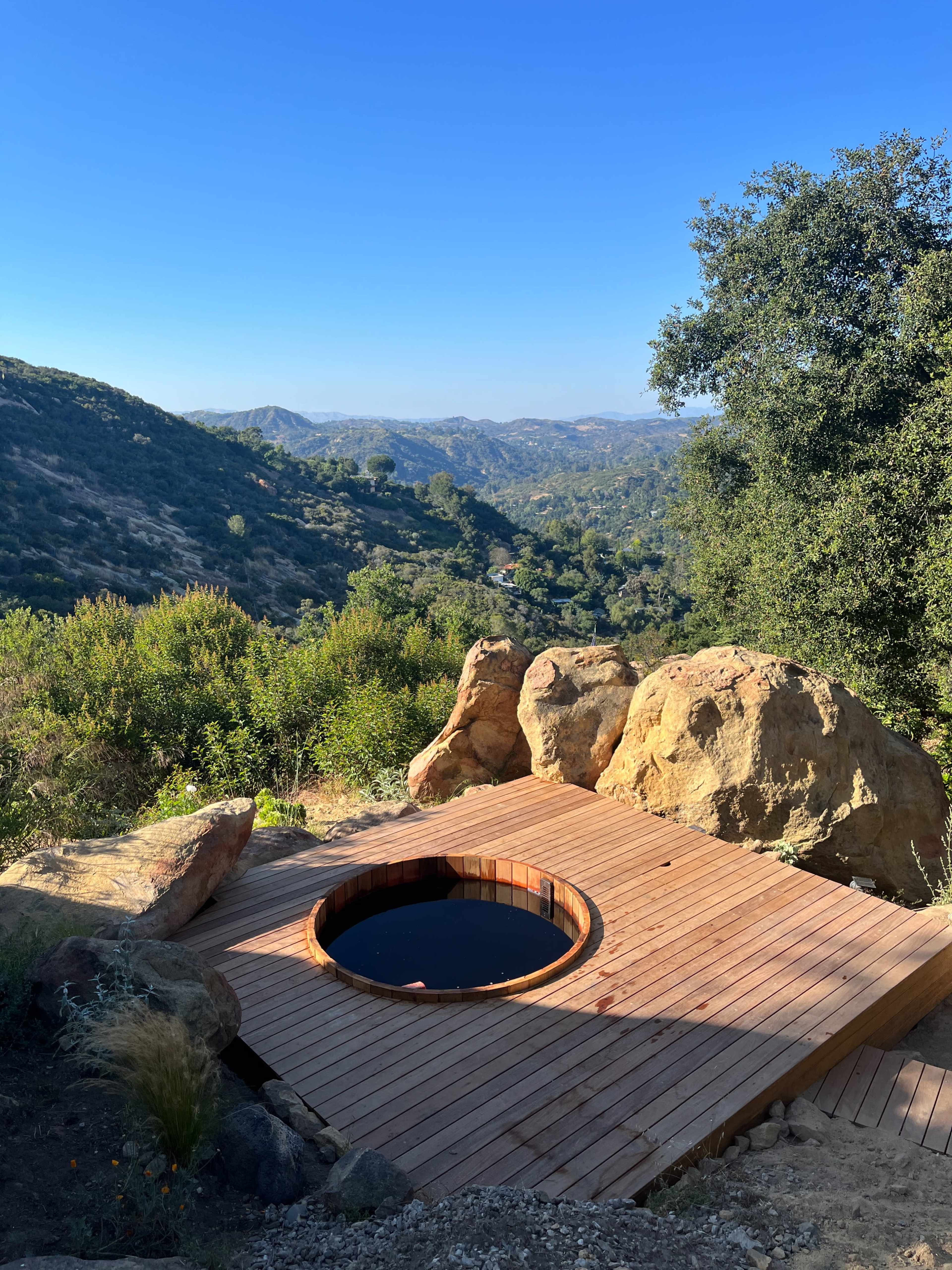 A circular wooden hot tub is situated on a deck overlooking a mountainous landscape under a clear blue sky.