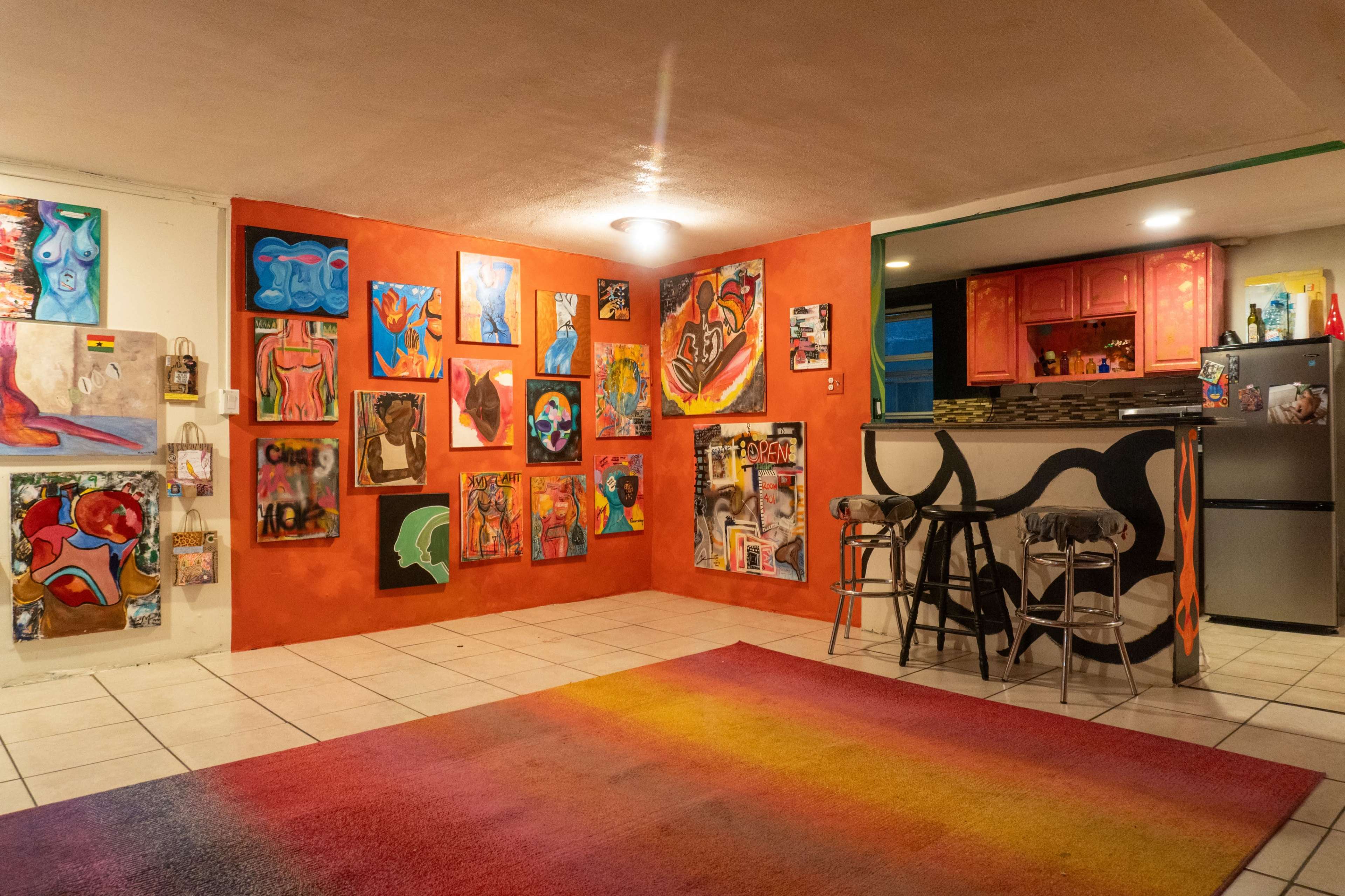 The image depicts a brightly colored room featuring an array of abstract paintings on one wall, while the opposite side shows a kitchen area with red cabinets and a bar with stools.