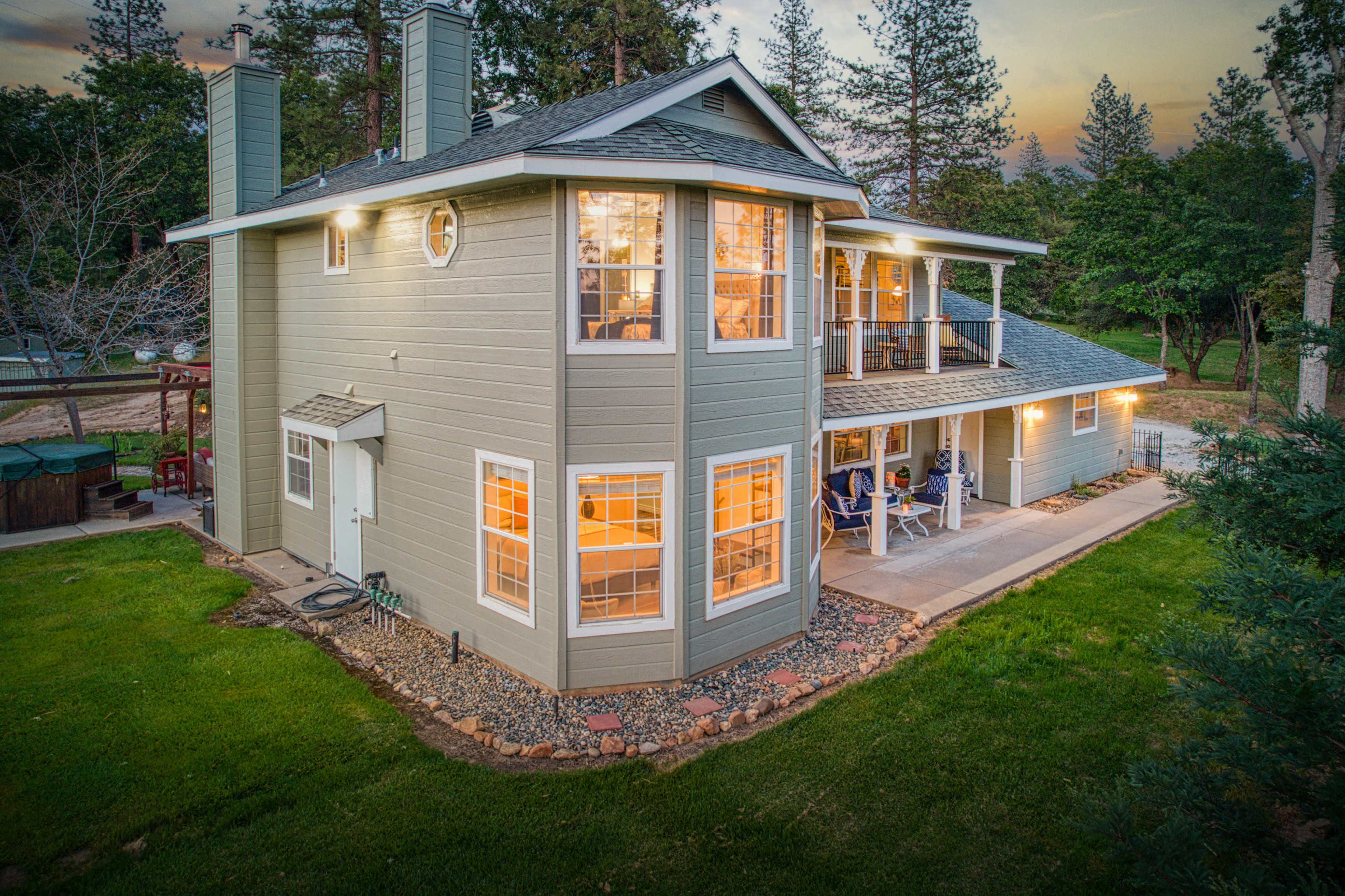 A two-story house with large windows, surrounded by greenery and a patio area, is illuminated in the evening light.