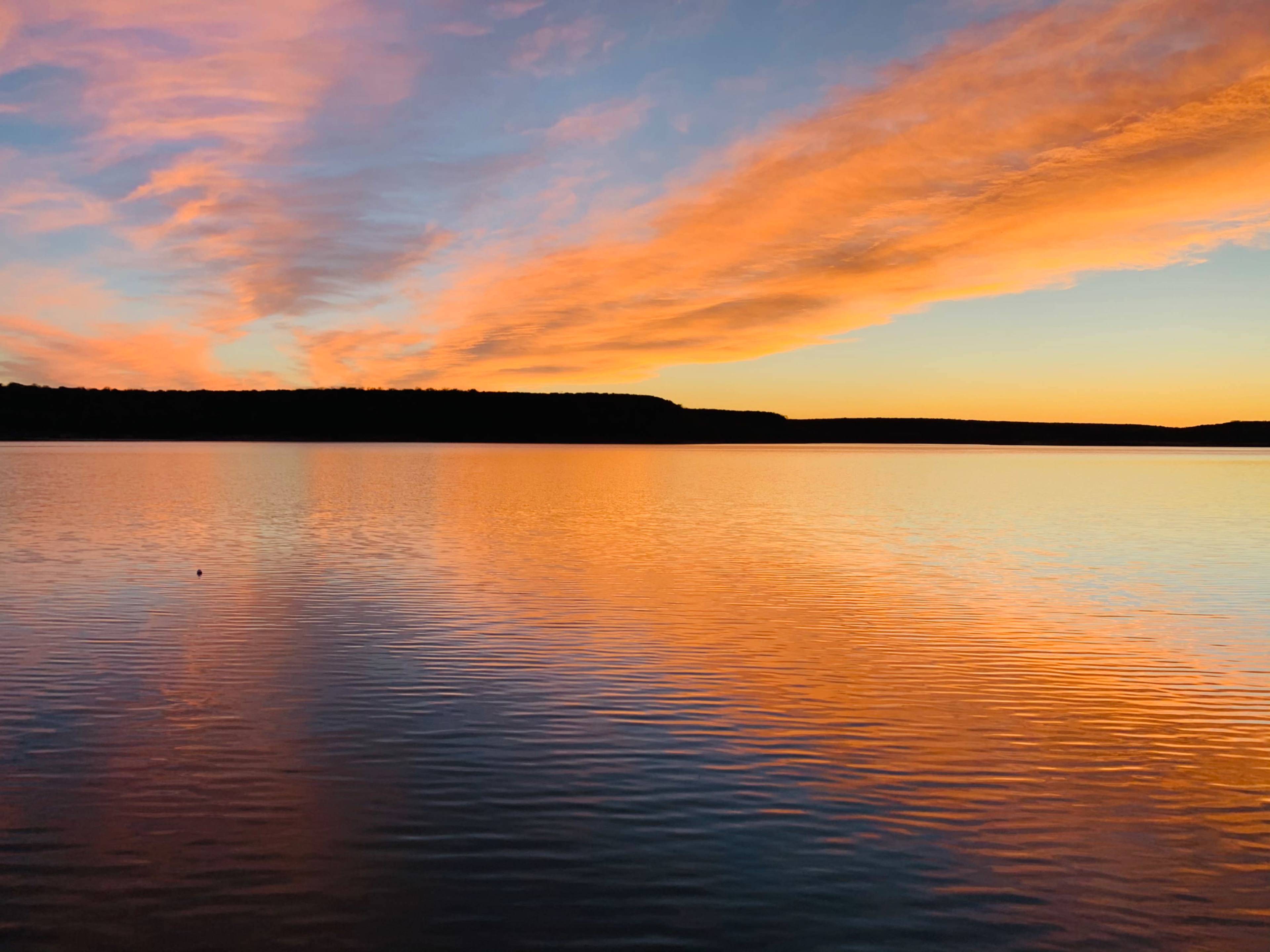 A tranquil lake reflects vibrant orange and pink clouds during sunset, framed by a distant tree line.