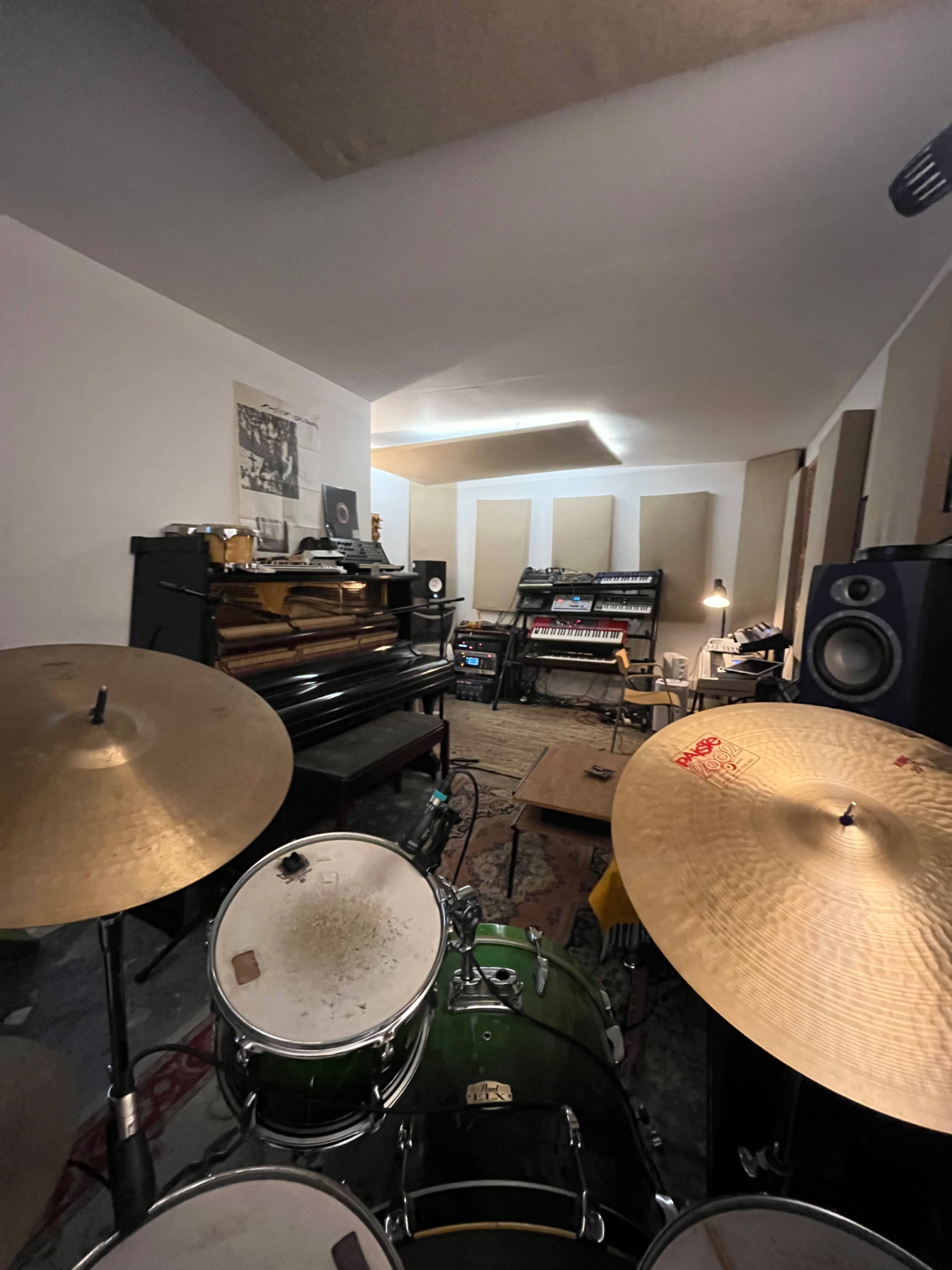 The image shows a music studio with a drum set in the foreground, a piano, and various electronic equipment arranged around the room.
