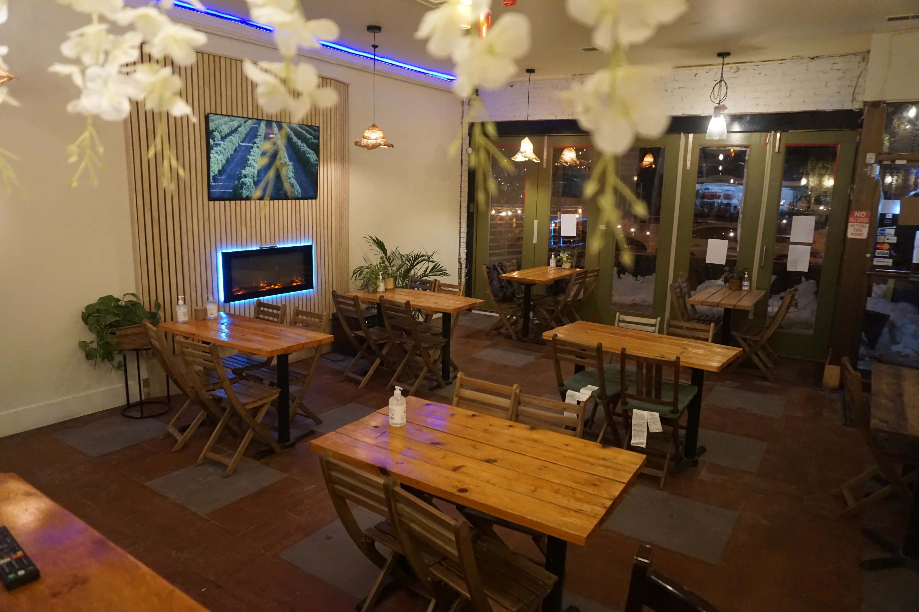 A dimly lit restaurant interior with wooden tables and chairs, a small electric fireplace, and plants near the windows.