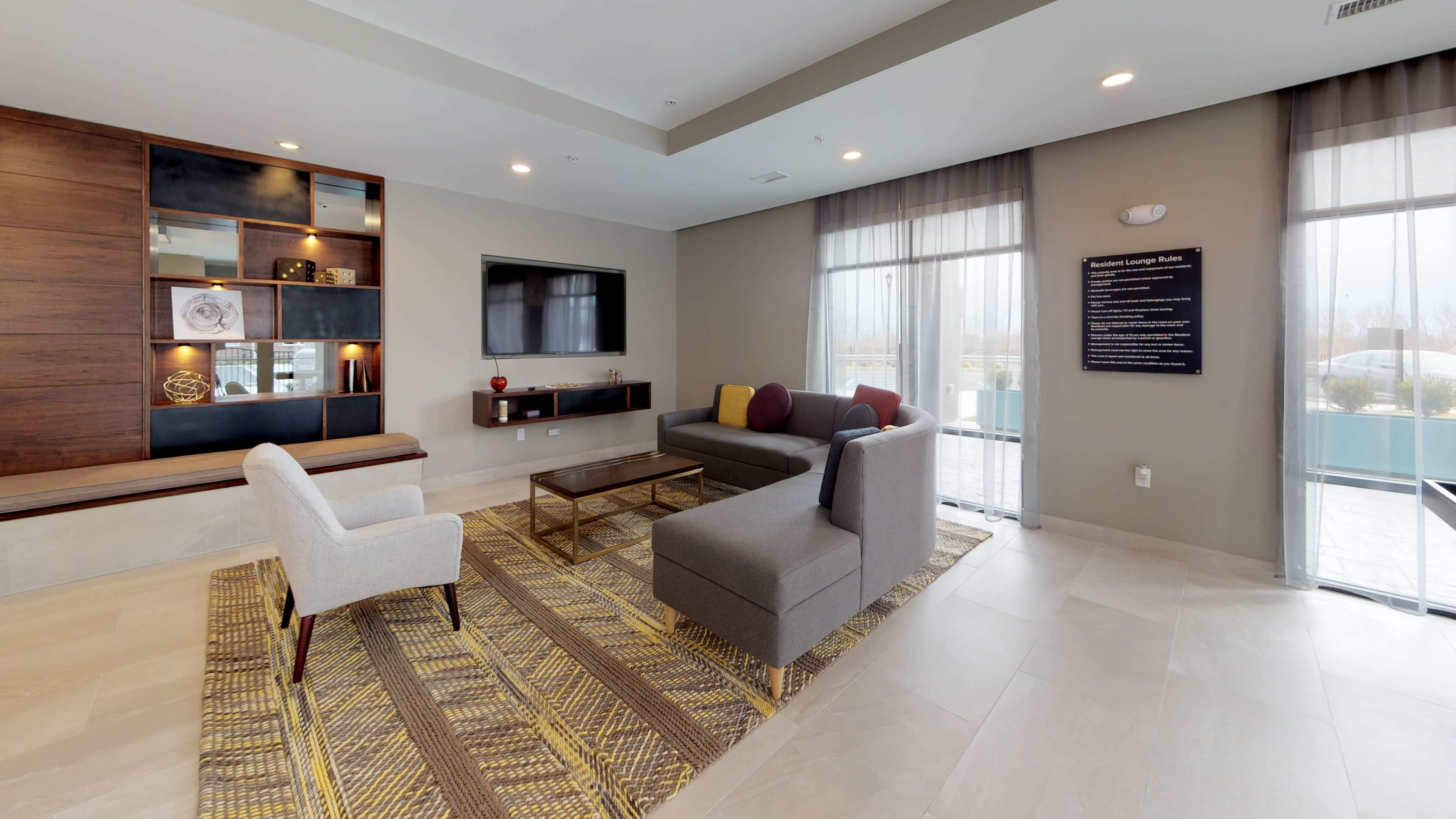 The image shows a modern lounge area featuring a sofa, a chair, a coffee table, and a wall-mounted television, with large windows allowing natural light to enter.