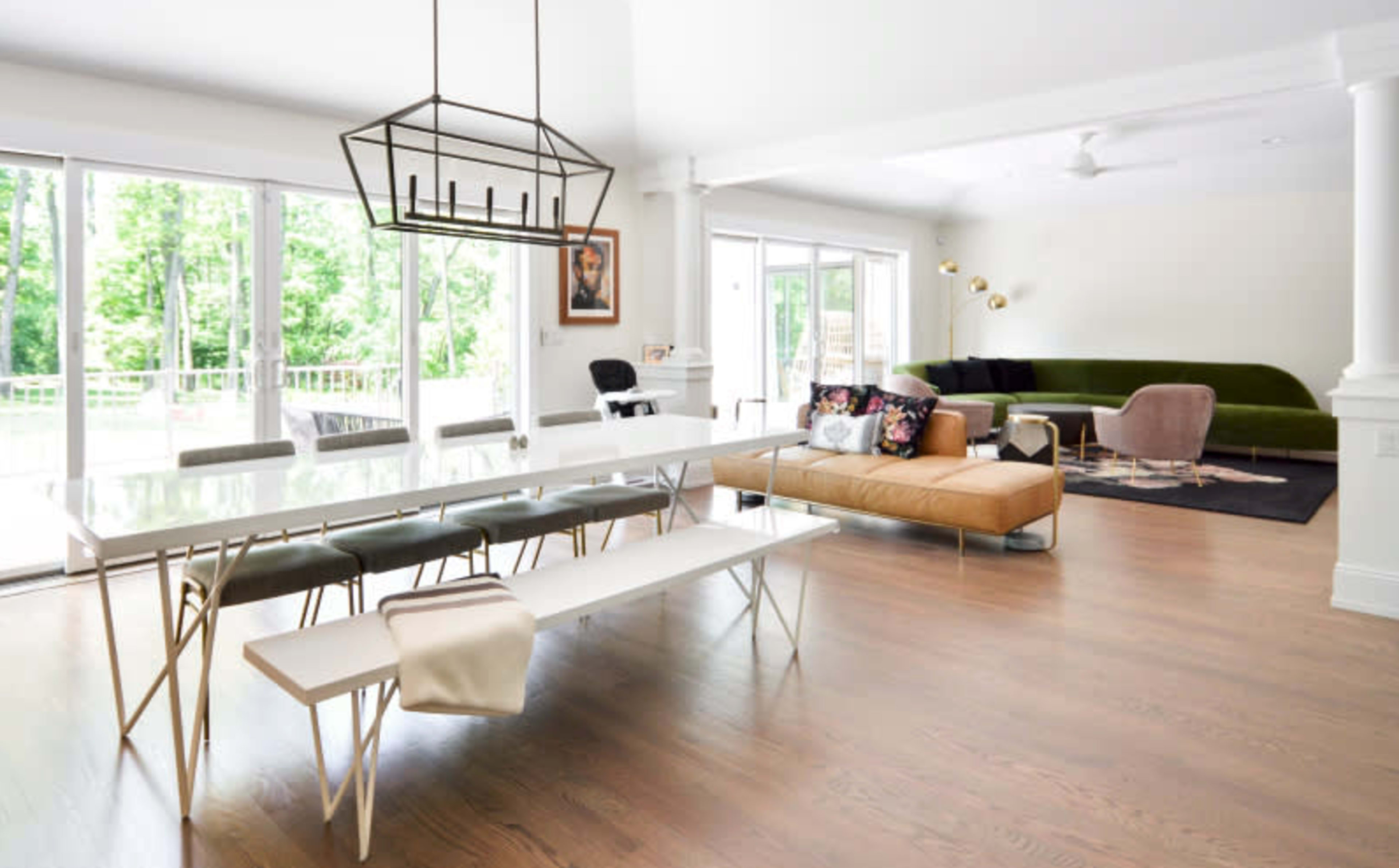 A modern living and dining area features a rectangular glass dining table with a bench and chairs, a sectional sofa, and large windows overlooking a green landscape.