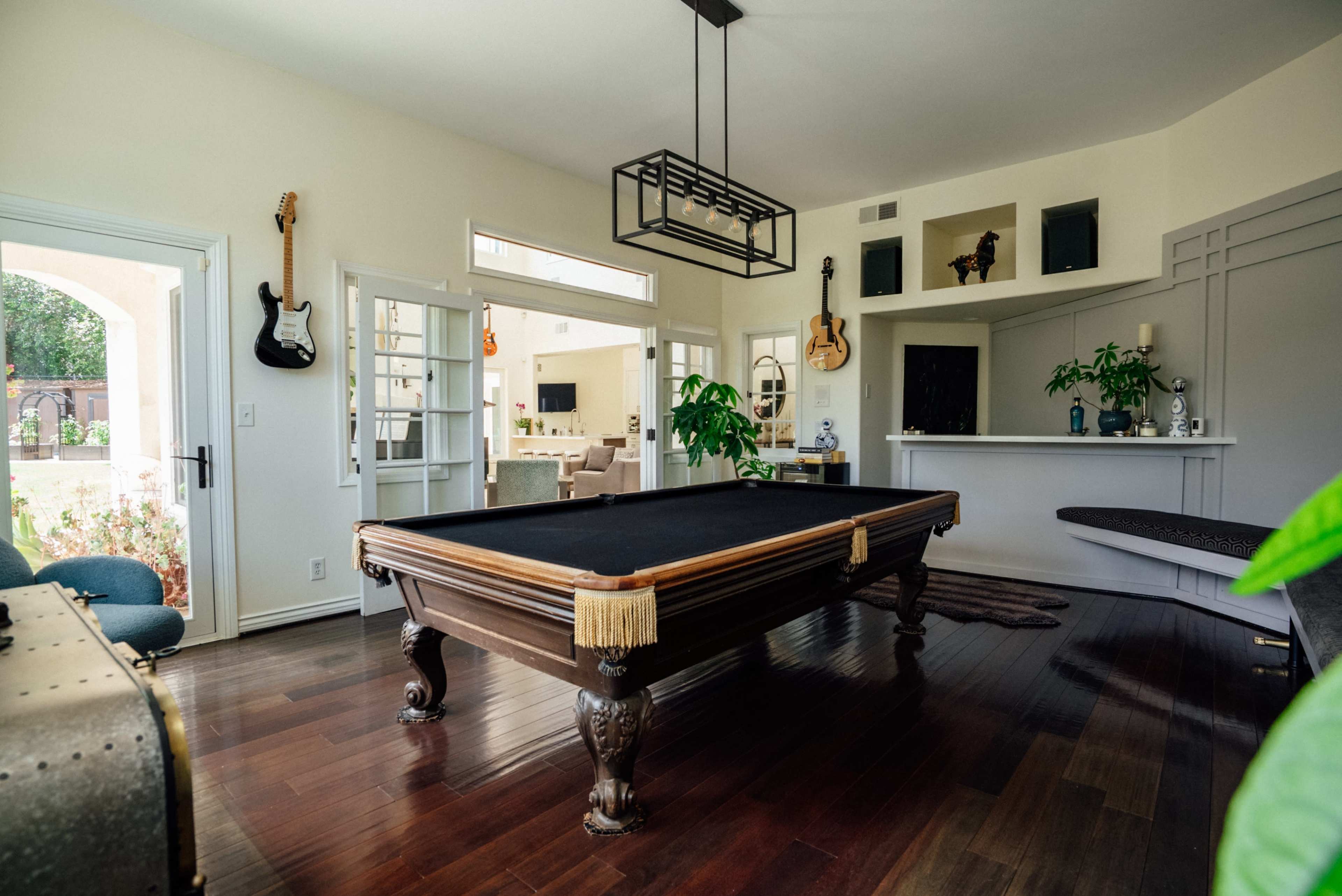 The image shows a spacious room featuring a pool table at the center, surrounded by large windows and light-colored walls with decorative musical instruments on display.