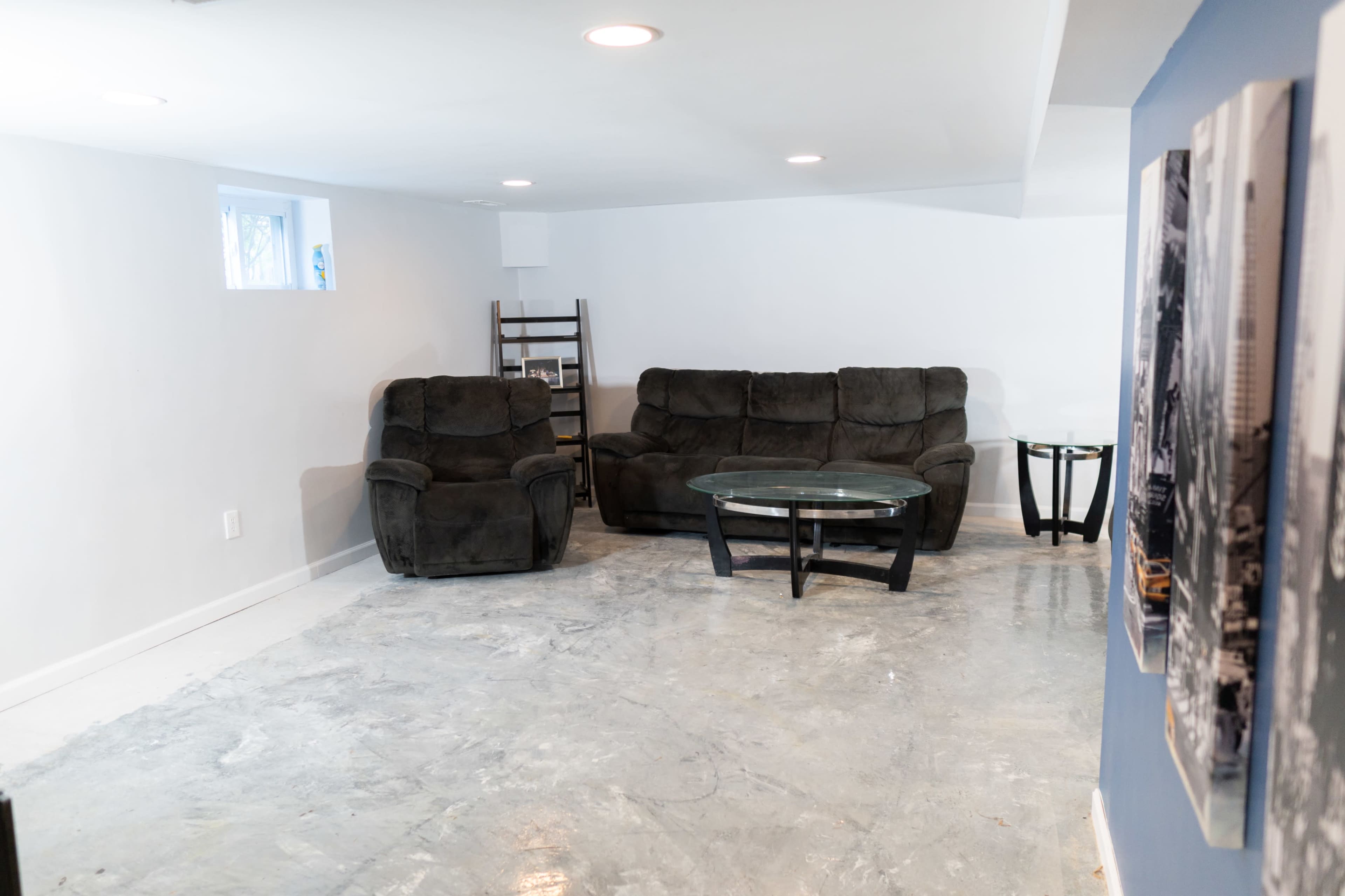 The image shows a sparsely furnished basement with two dark brown couches and a glass coffee table on a concrete floor.