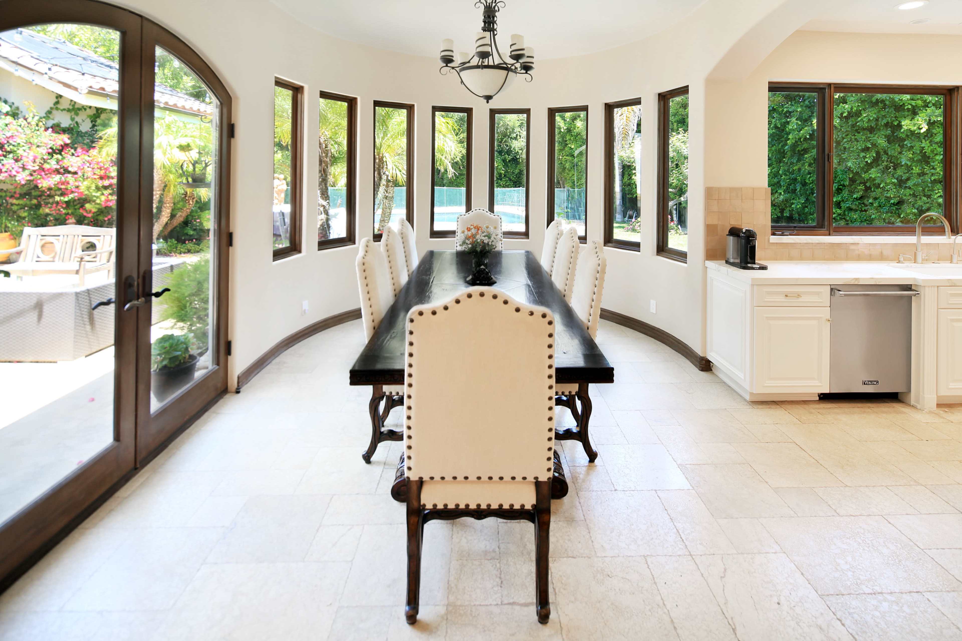 A spacious dining area features a long black table surrounded by twelve light-colored chairs and large windows offering views of a lush outdoor landscape.