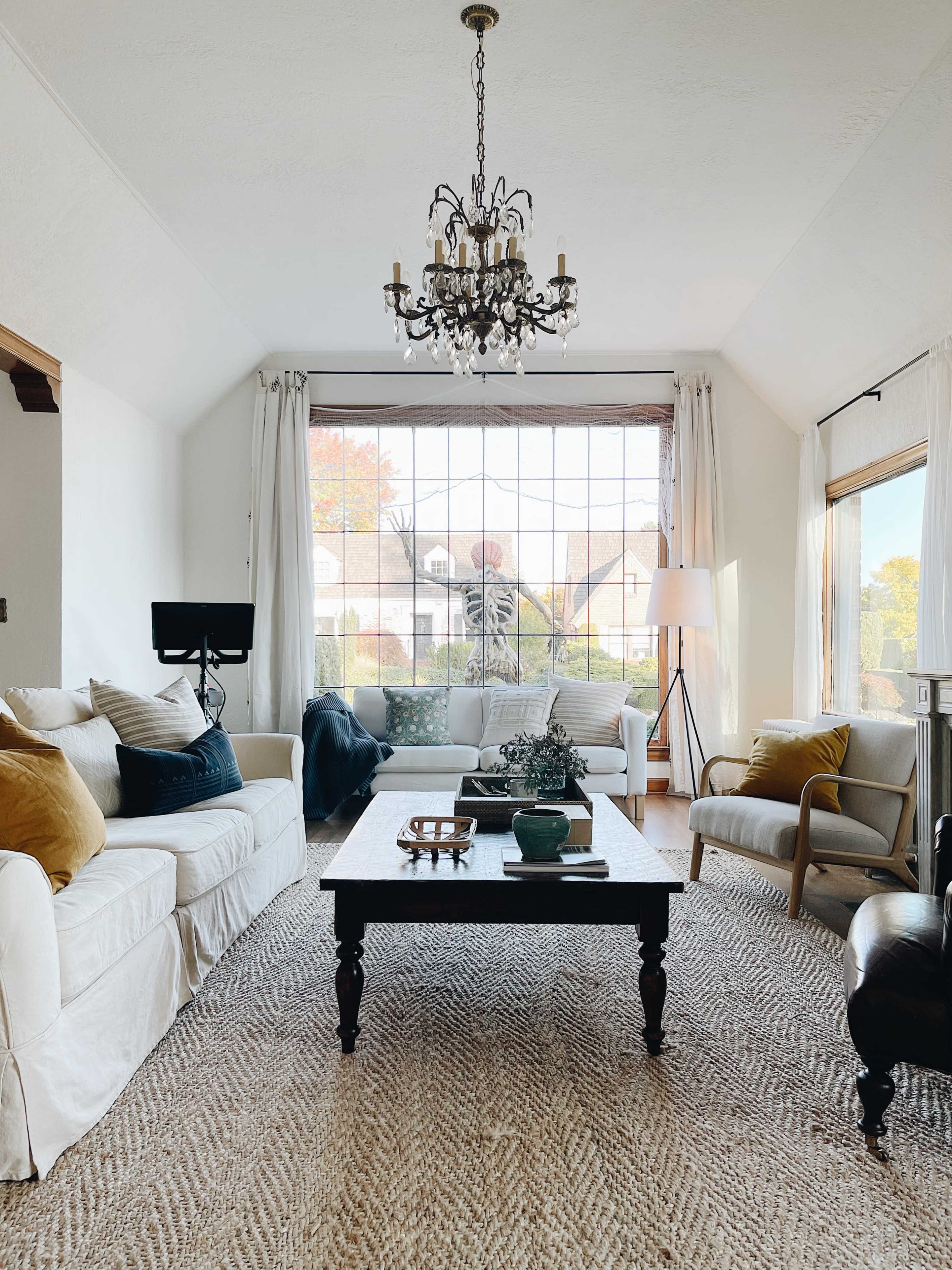 A bright living room features two white sofas, a black coffee table, a woven rug, and large windows with a view of a skeleton outside.