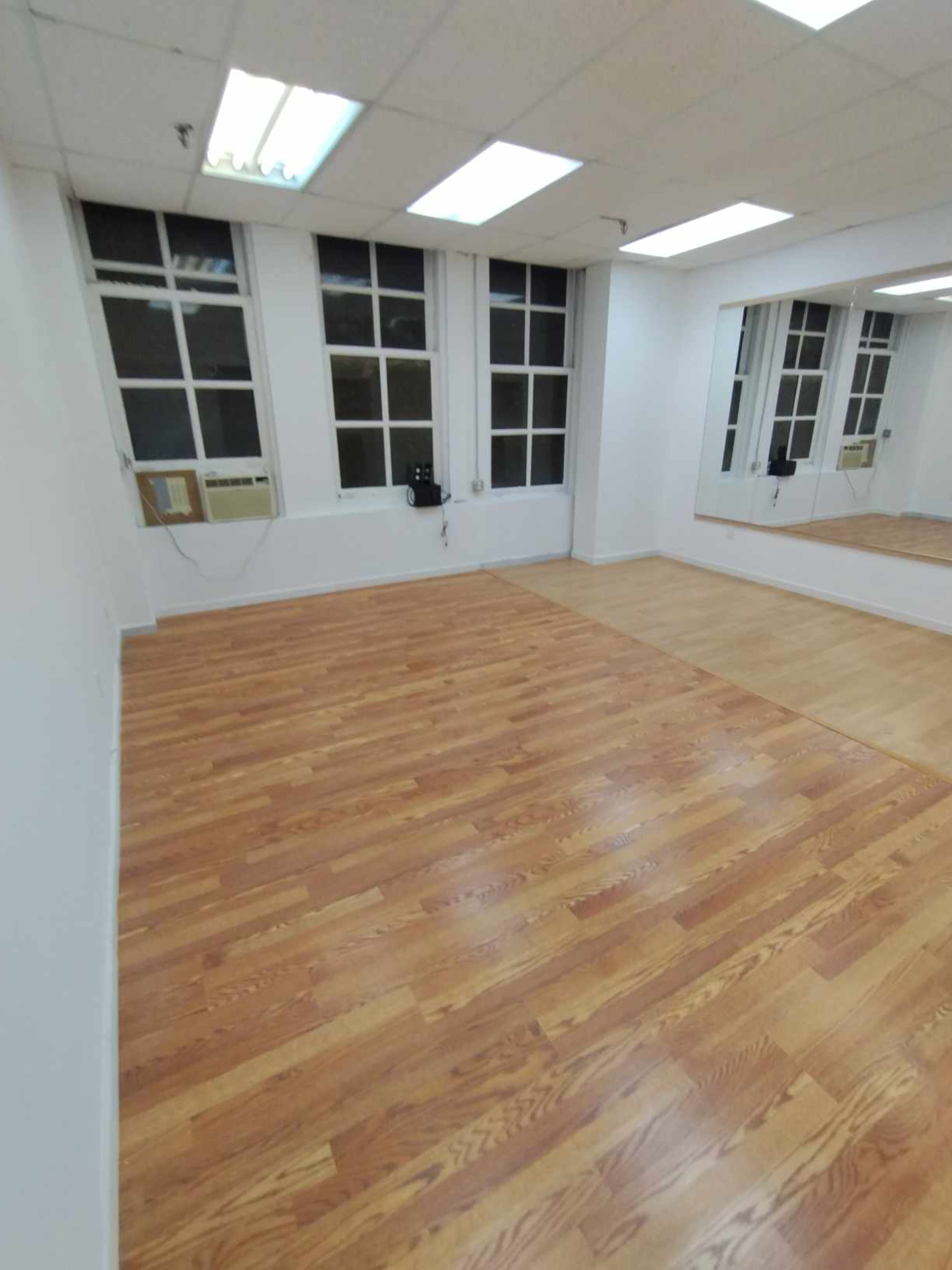 The image shows a spacious room with wooden flooring, large windows on one side, and a mirror mounted on the opposite wall.