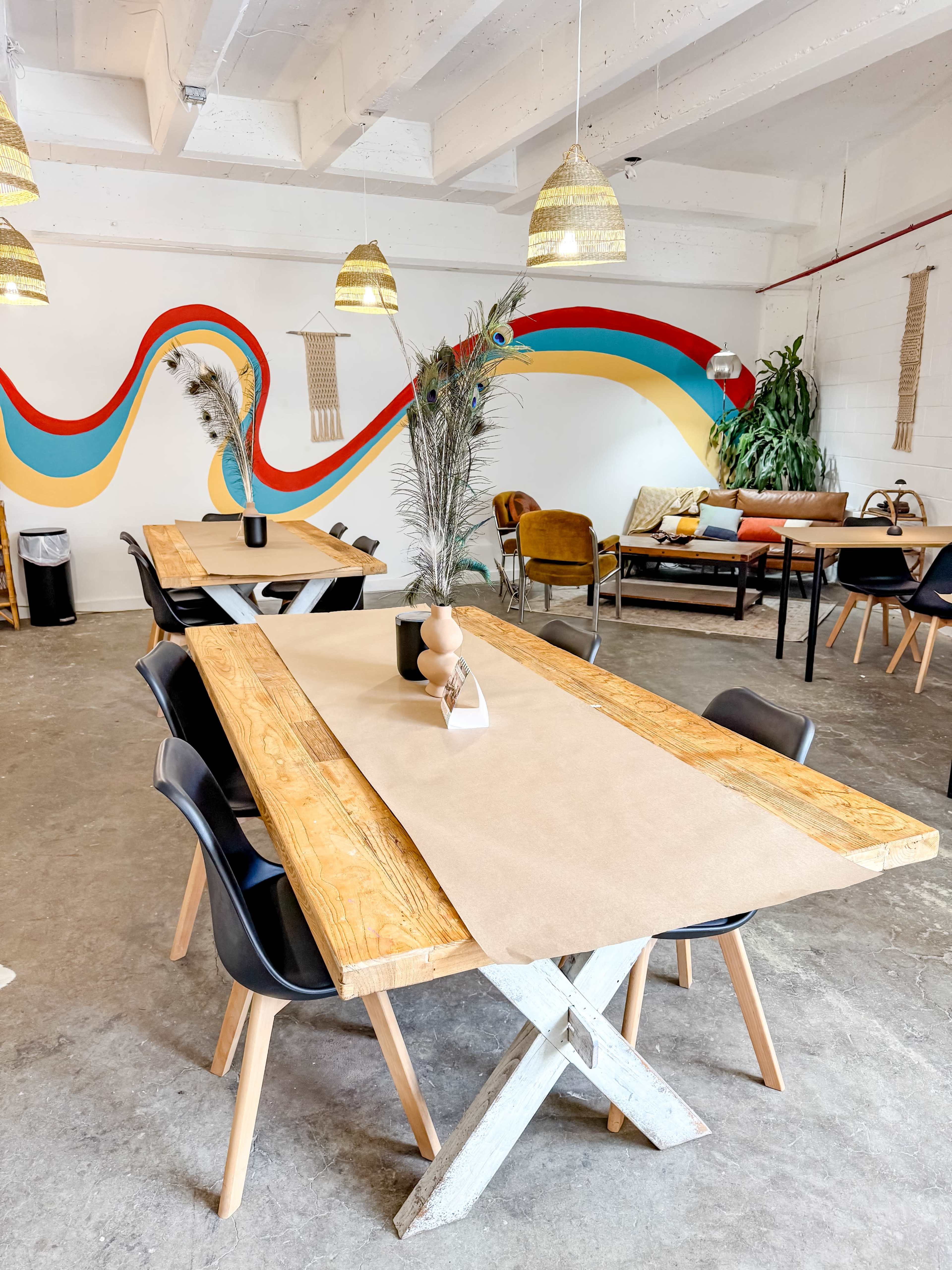 The image shows a bright, modern café space featuring wooden tables, black chairs, and colorful wall decorations.