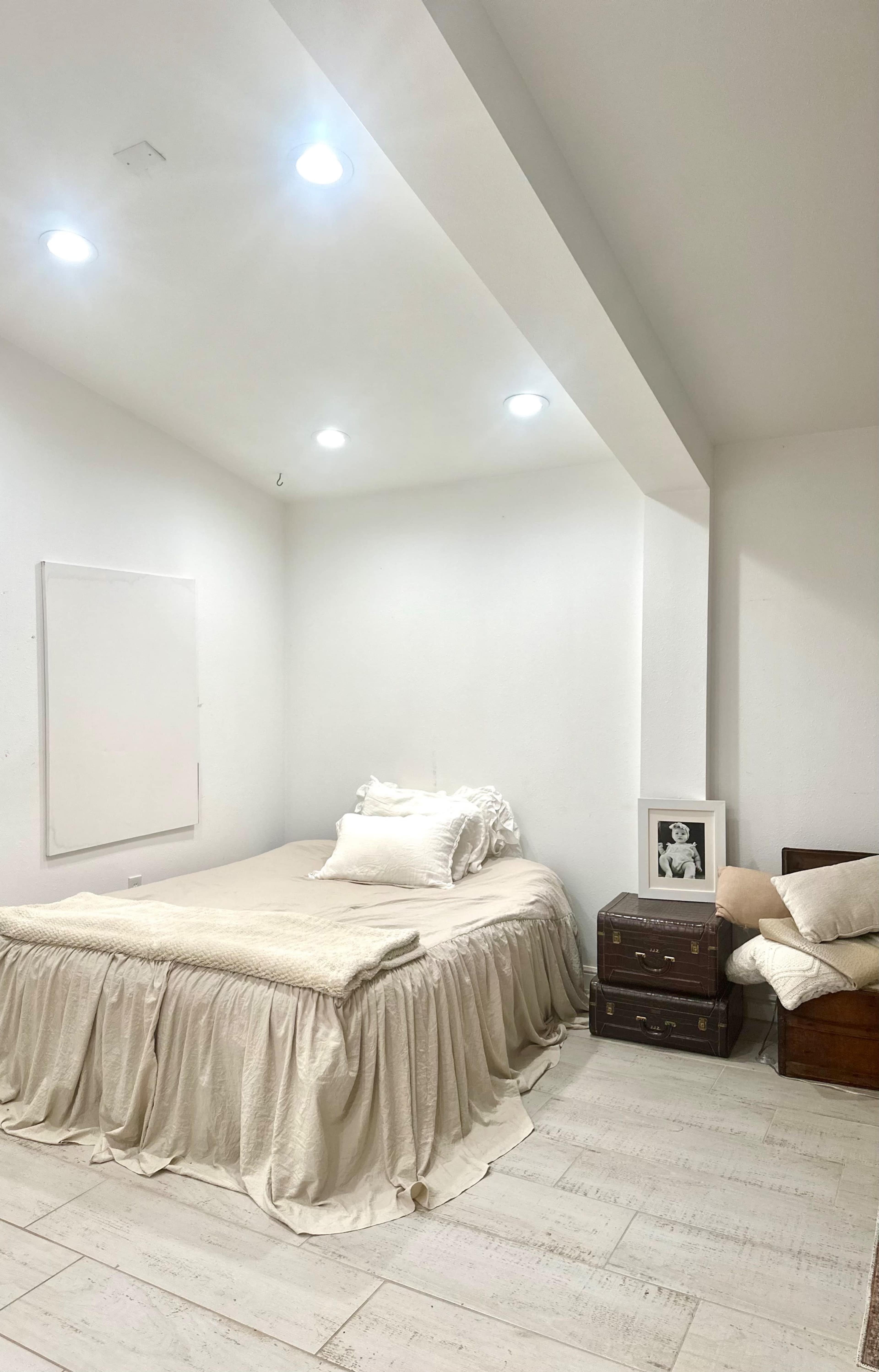 A minimalistic bedroom with a bed covered in a light-colored blanket, a wooden nightstand, and a framed photo on the floor beside a small couch.