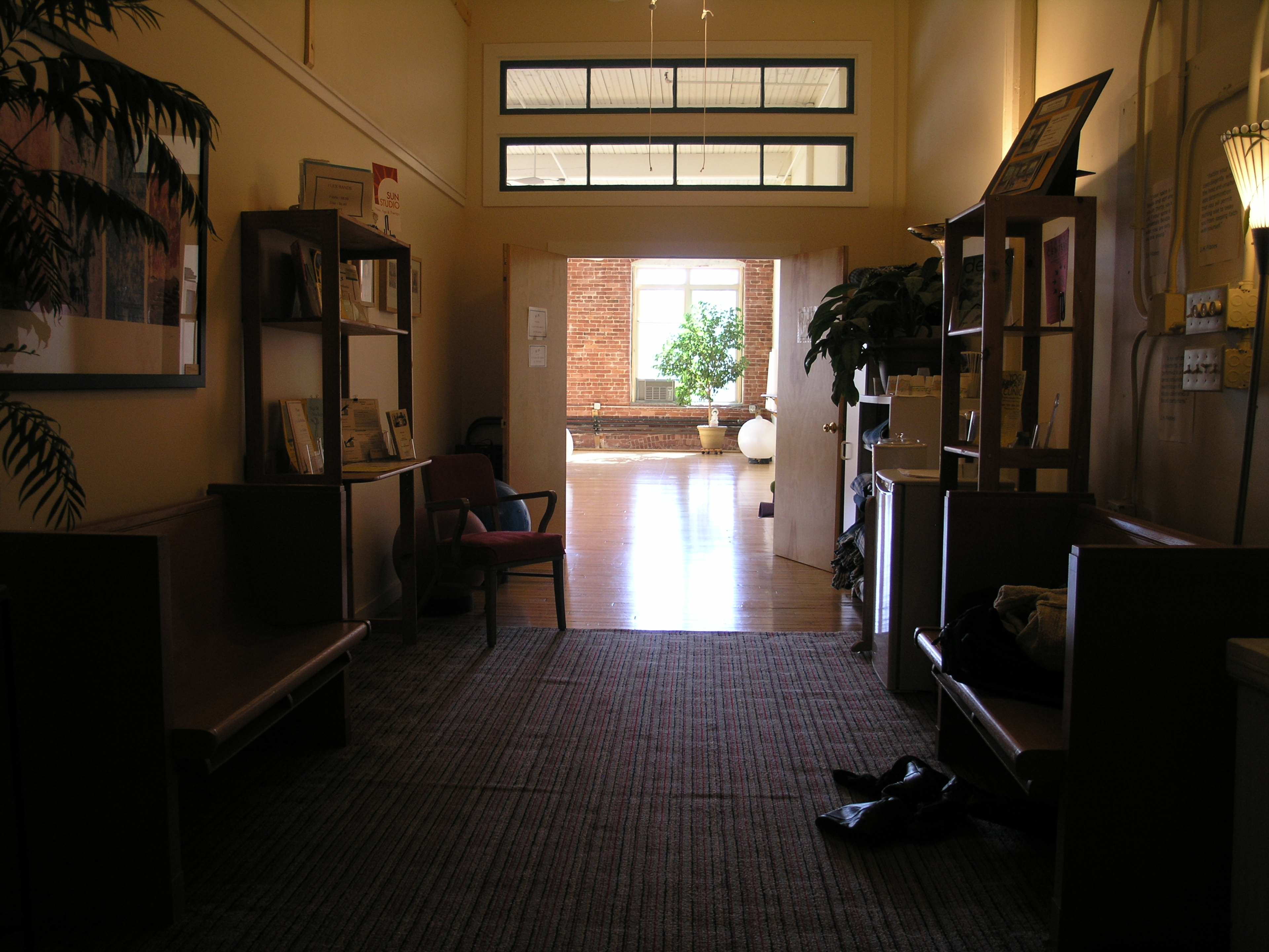 A hallway leading to a room with large windows and plants, flanked by wooden benches and shelves filled with books and decor.