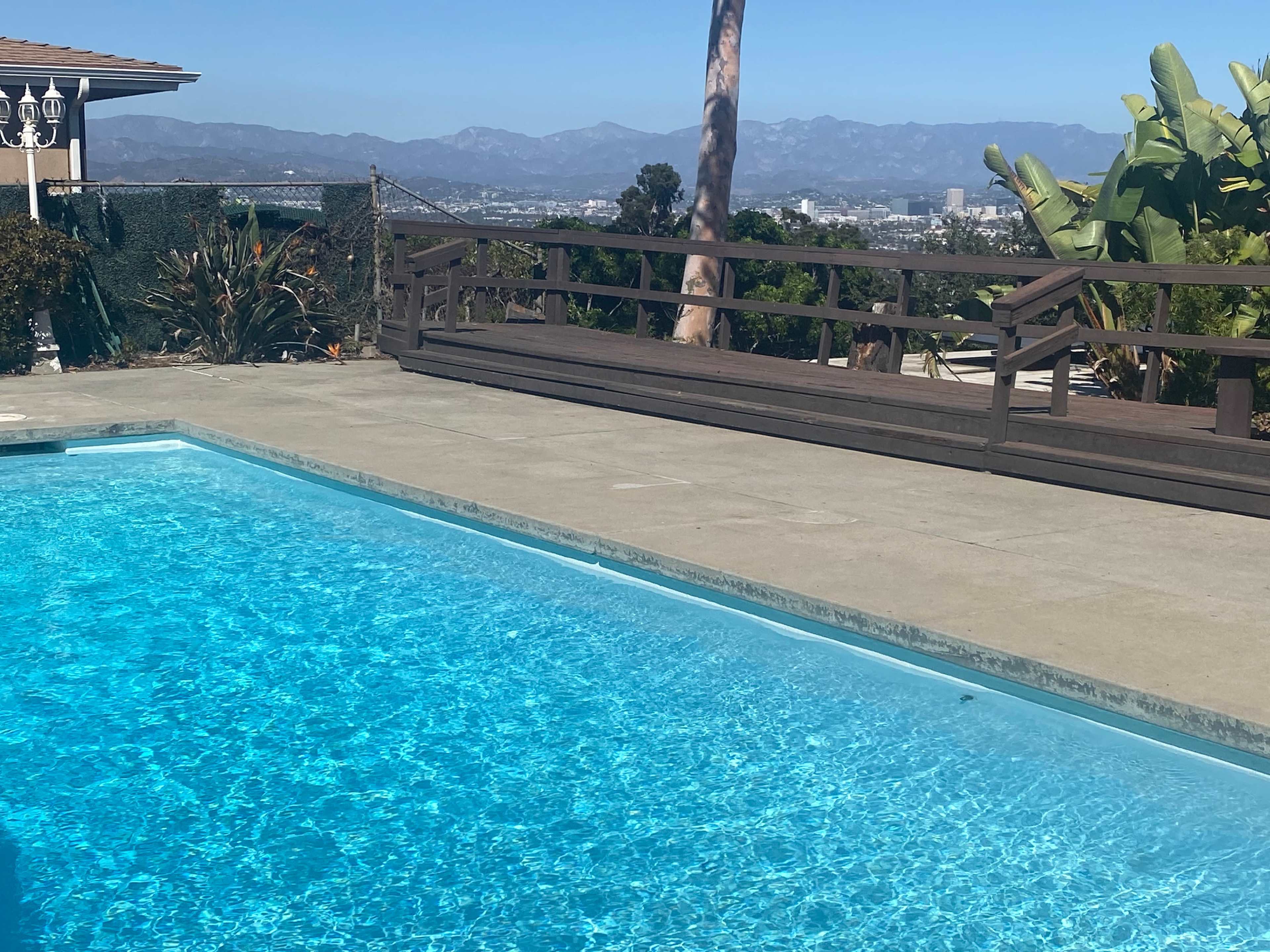 A clear swimming pool beside a wooden deck with a view of mountains and a city in the background.